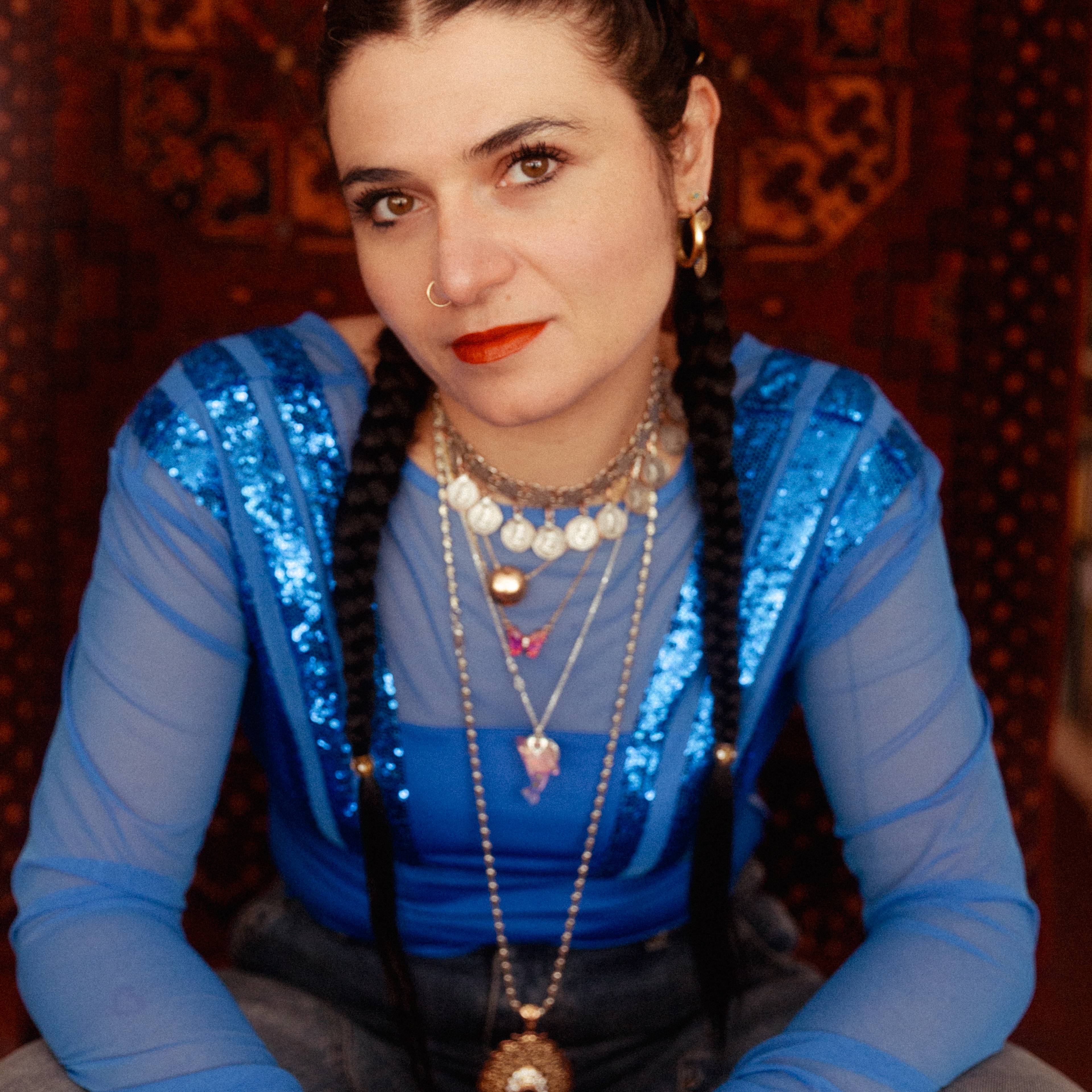 A person with long dark hair styled in braids is seated in front of a patterned background, wearing a sheer blue top with sequins, layered necklaces, and red lipstick.