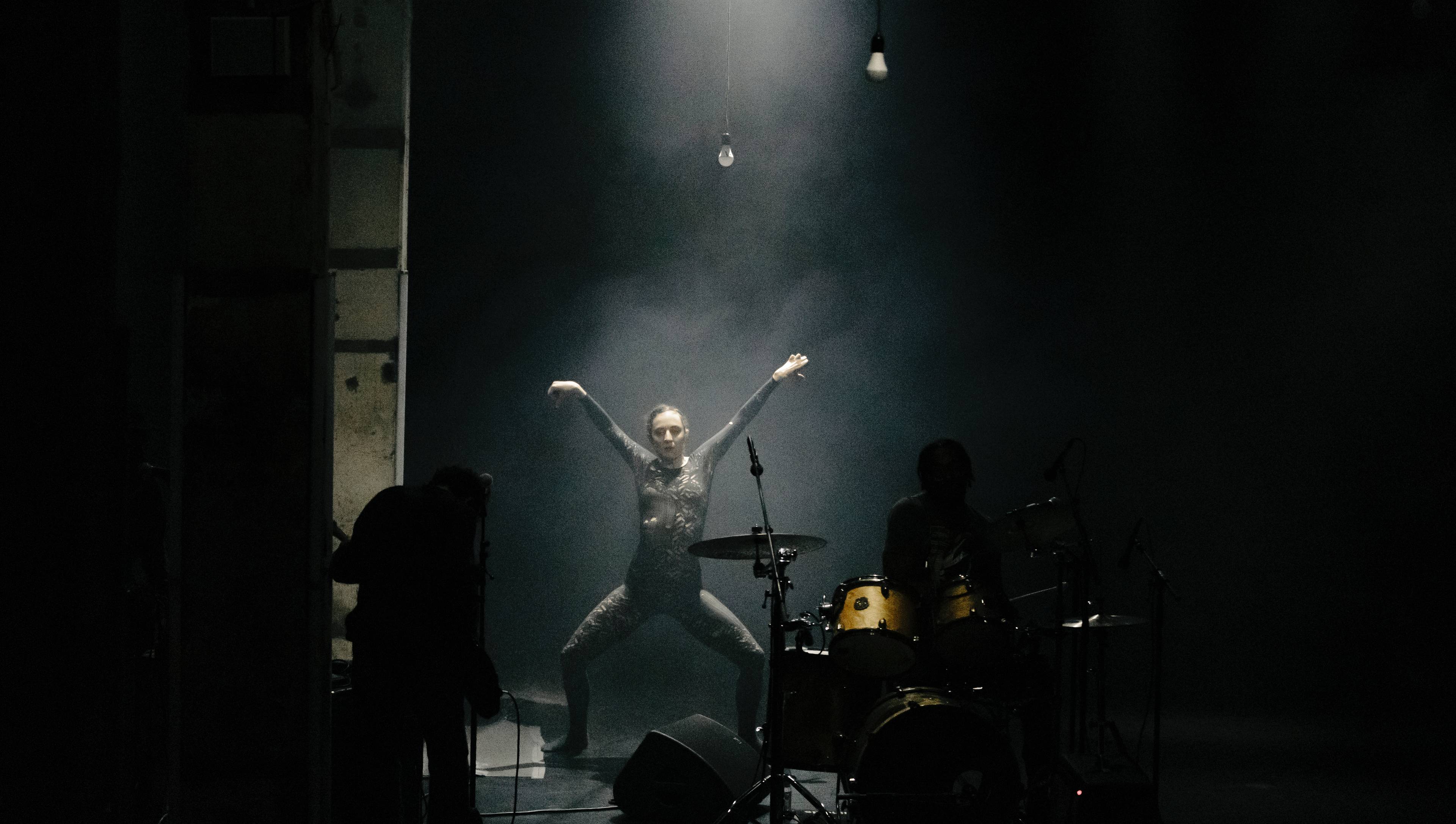 Wide-angle photograph of a woman dancer with legs and arms splayed out in an X shape, lit from above by a spotlight.