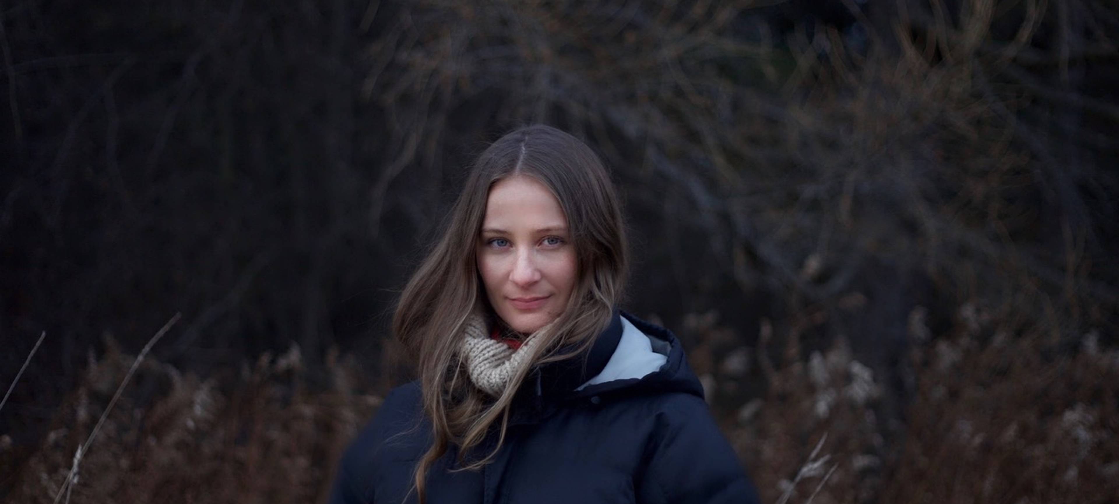 A person with long hair, a warm jacket, and a scarf stands in autumnal nature, gazing calmly into the camera.
