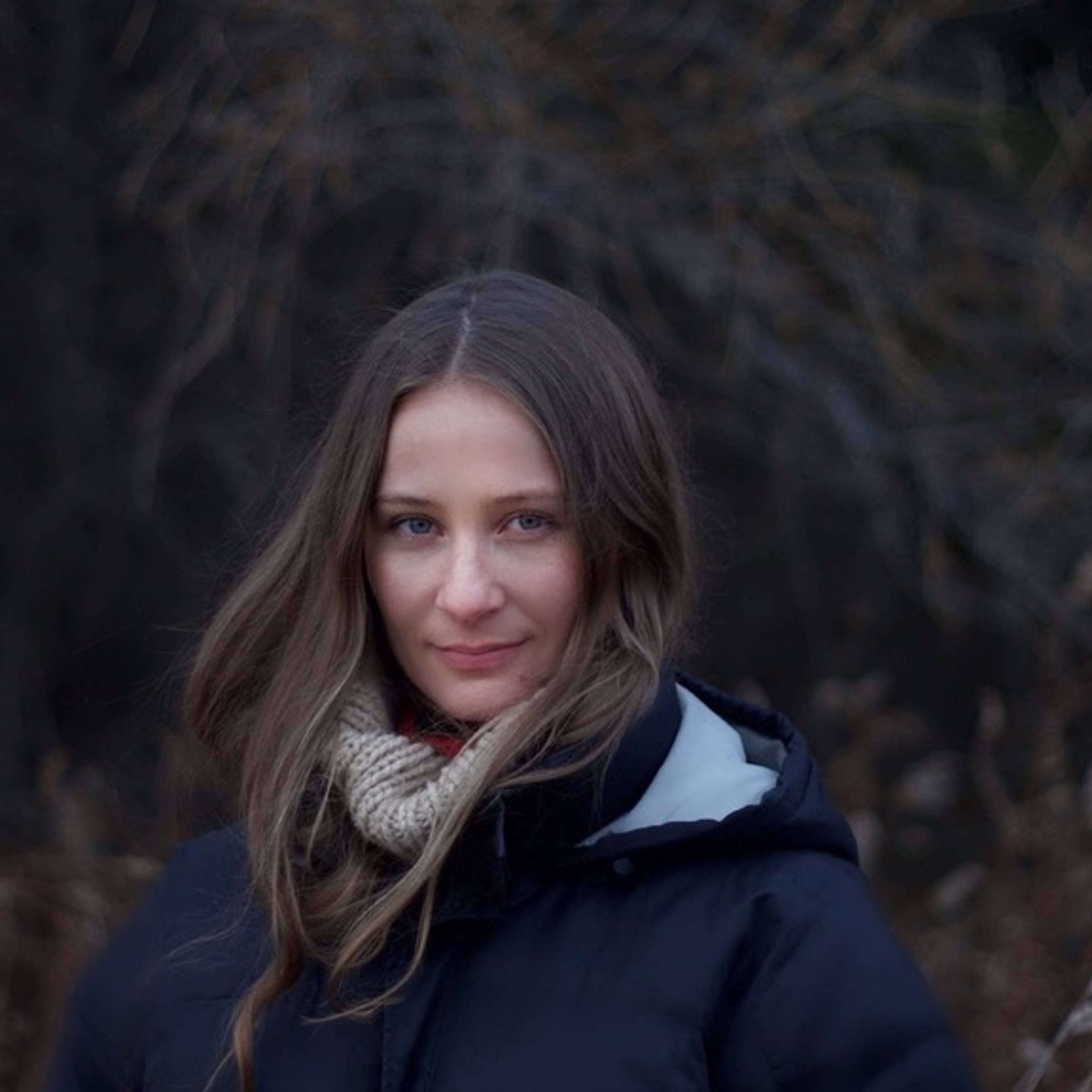 Eine Person mit langen Haaren, warmer Jacke und Schal steht in herbstlicher Natur und blickt ruhig in die Kamera.