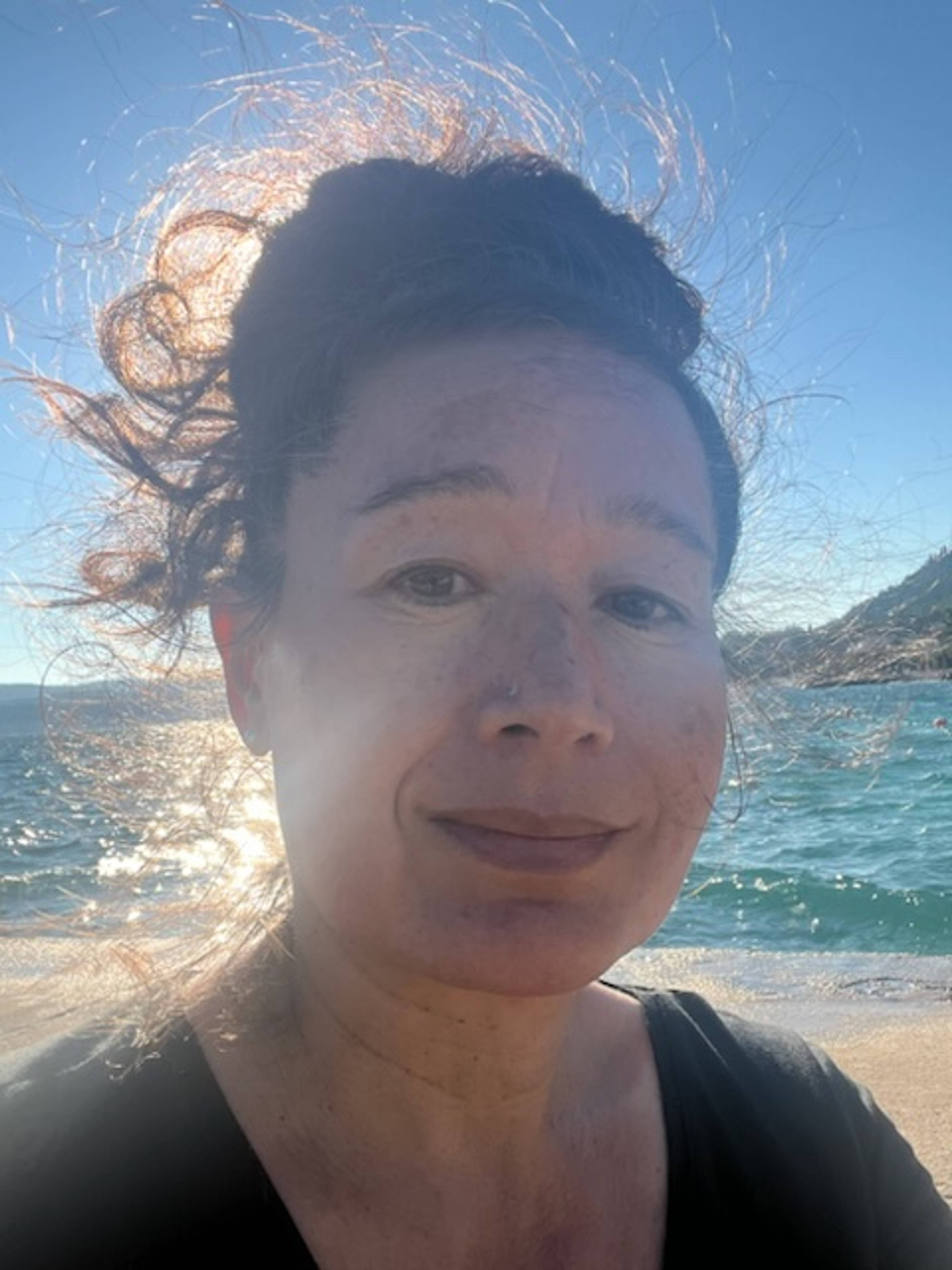 A person with curly hair tied up is outdoors near the sea, sunlight reflecting on the water behind them, smiling slightly at the camera.