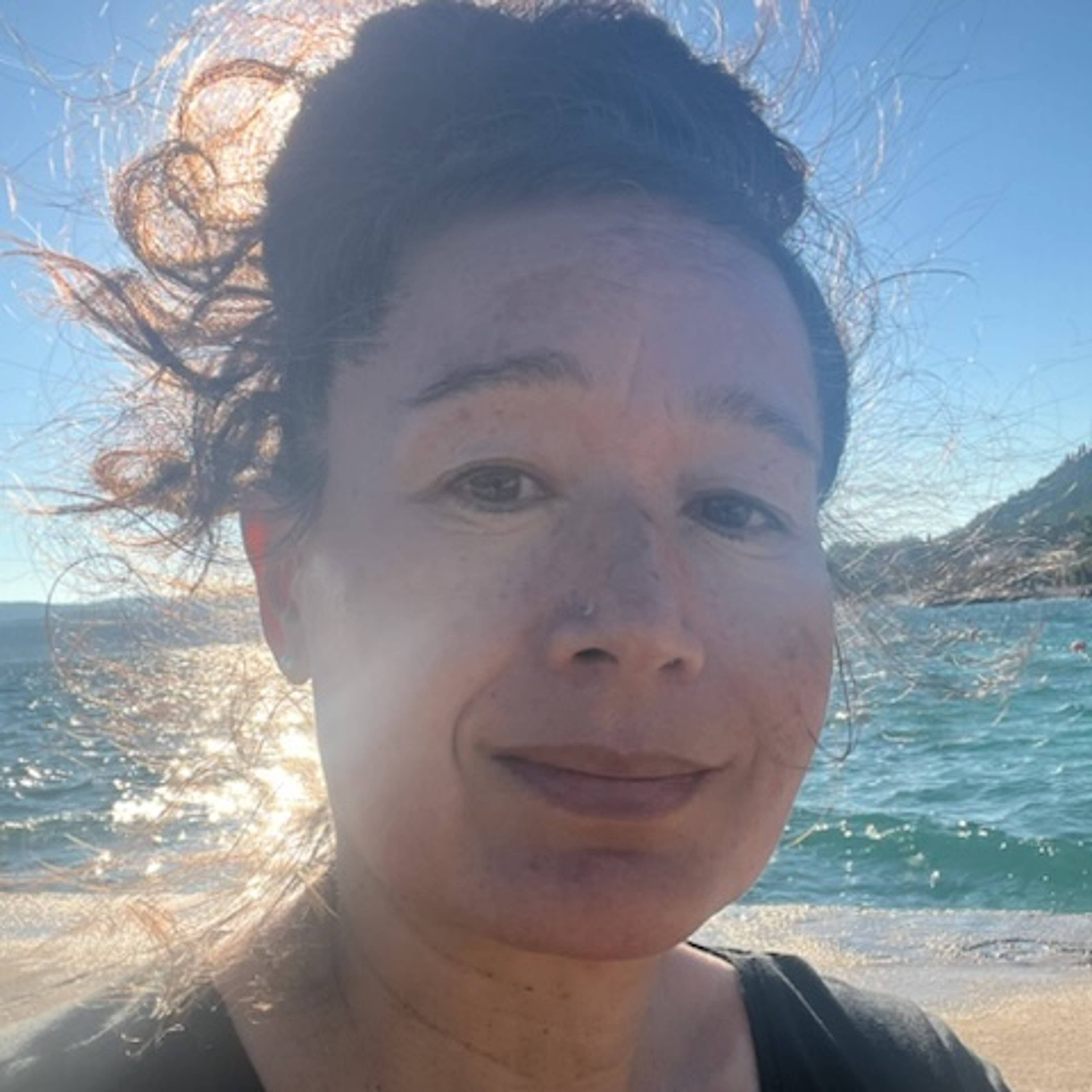 A person with curly hair tied up is outdoors near the sea, sunlight reflecting on the water behind them, smiling slightly at the camera.