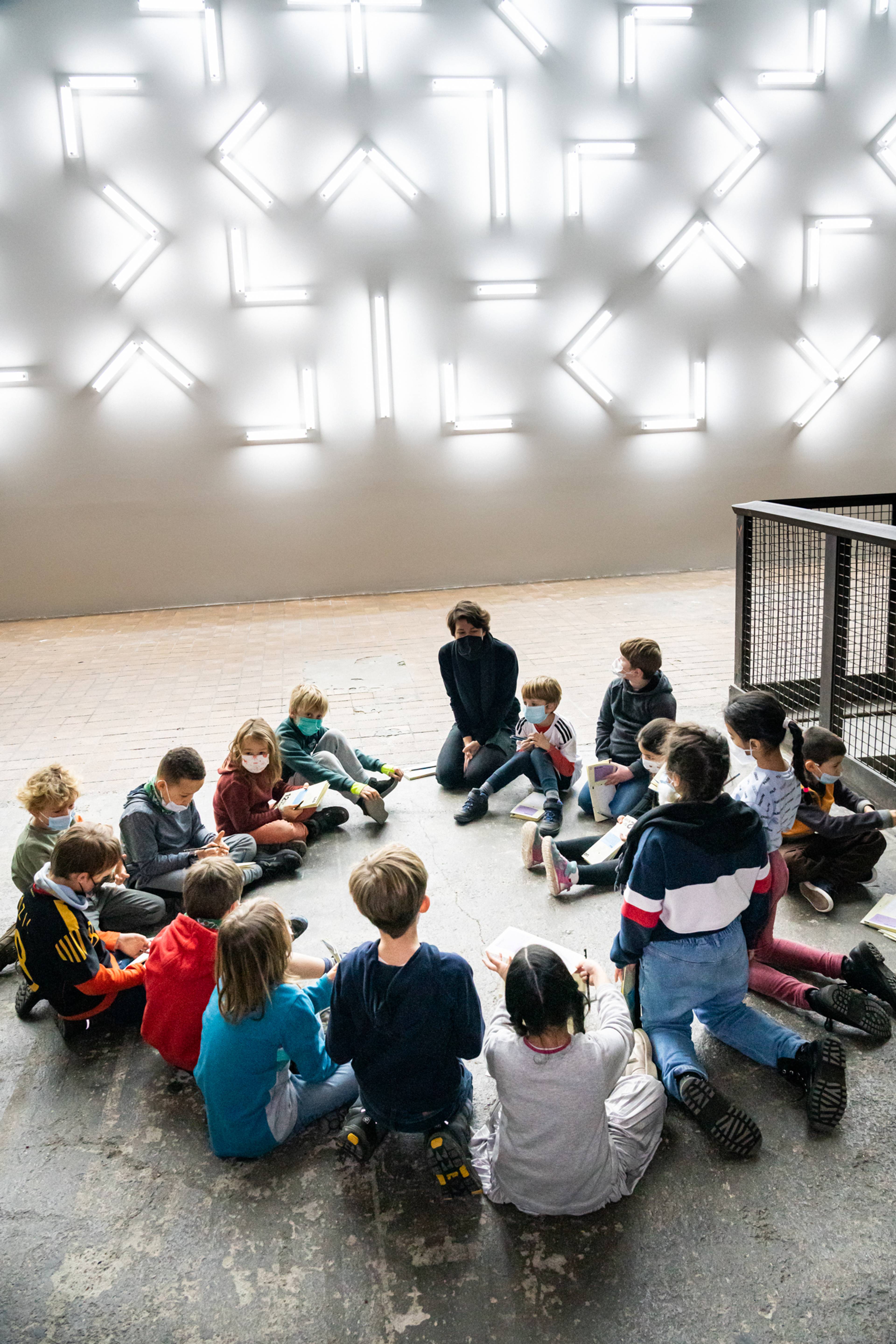 Eine Kunstpädagogin erklärt einer Gruppe von Schulkindern, die sich im Kreis auf dem Boden versammelt haben, die Lichtinstallation von Robert Irwin.