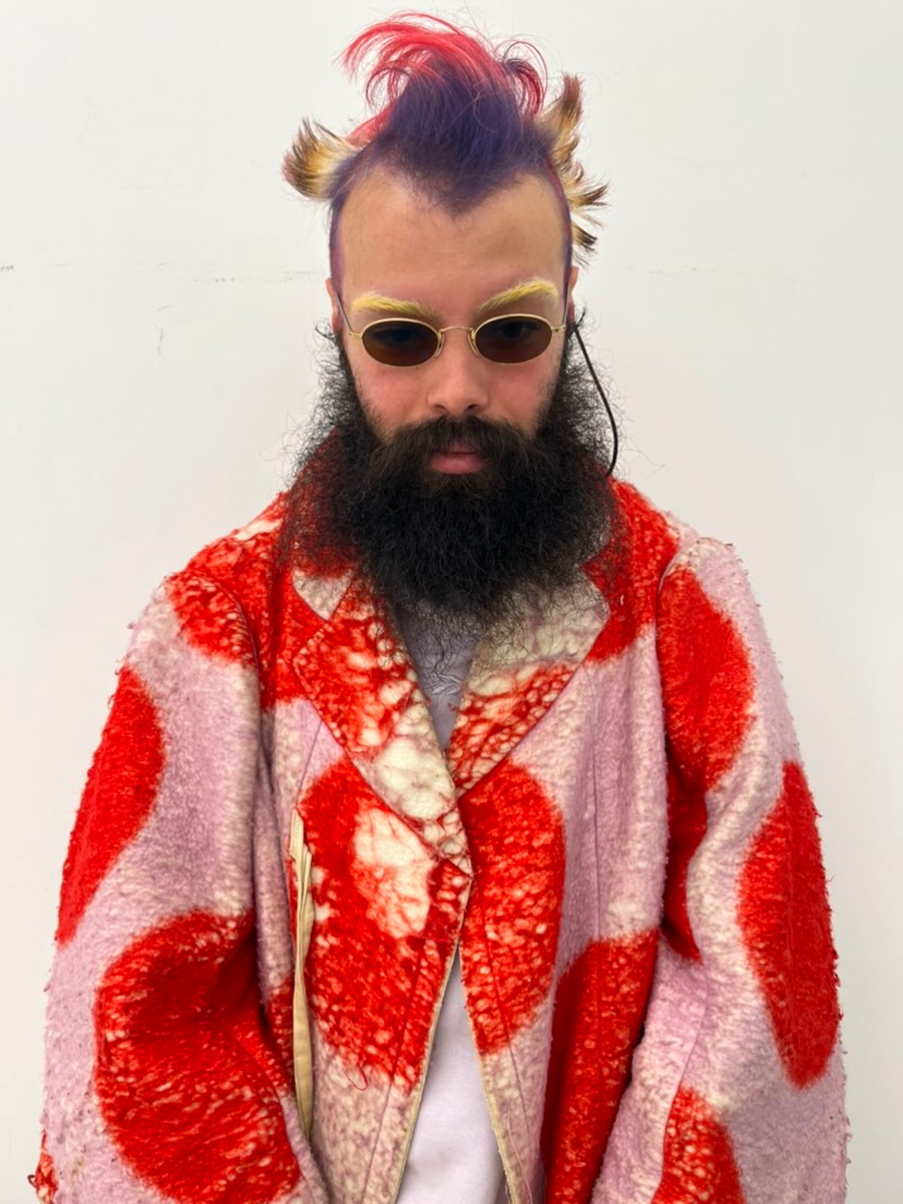 A person with colorful, spiked hair and a full beard wears cat-eye sunglasses and a pink coat with red circular patterns, standing against a plain white background.