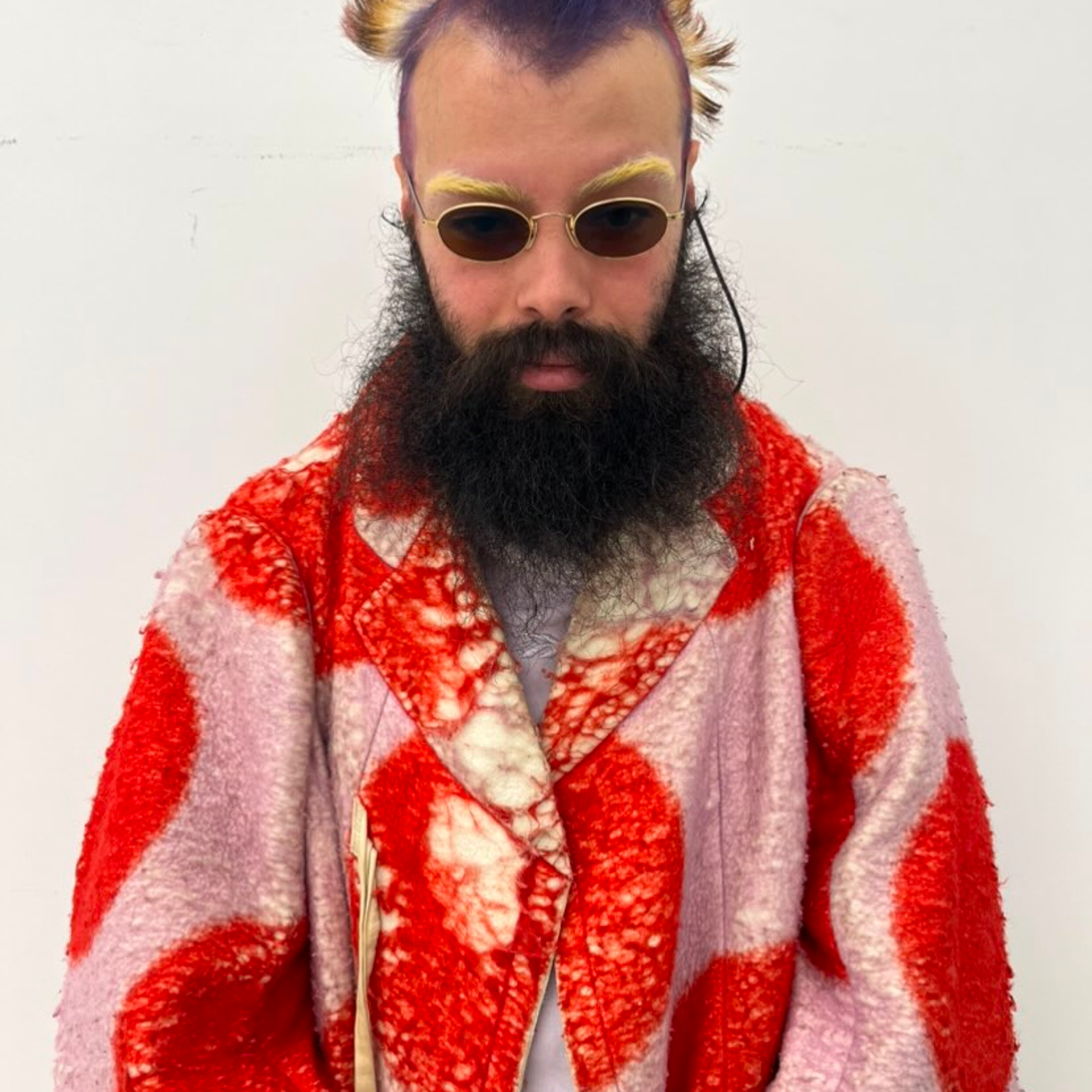 A person with colorful, spiked hair and a full beard wears cat-eye sunglasses and a pink coat with red circular patterns, standing against a plain white background.