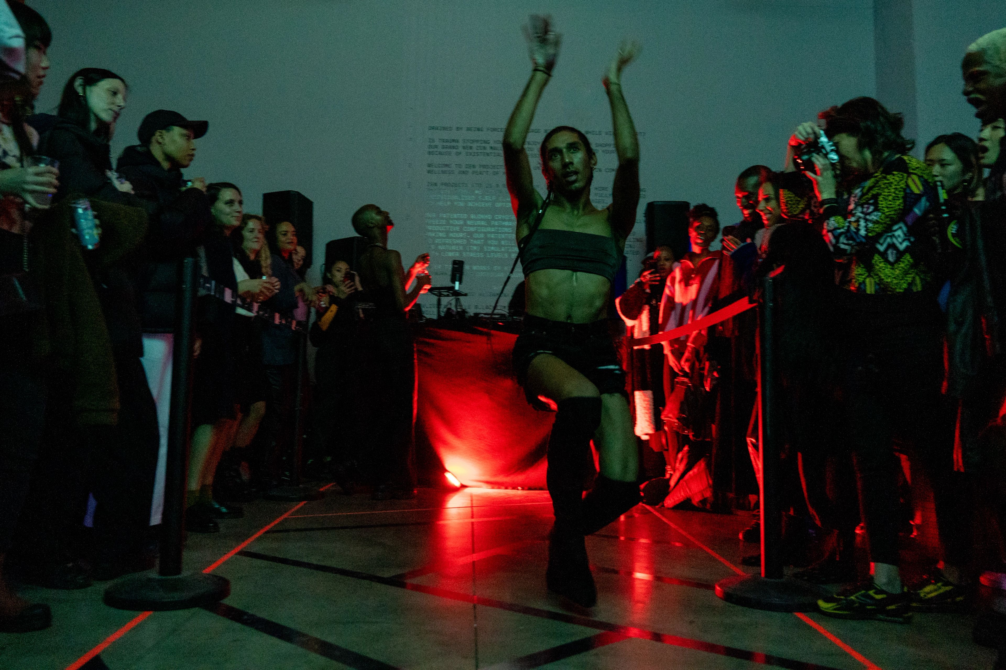 Ein:e Performer:in posiert während eines Tanzes in einem schwach beleuchteten Raum, umgeben von einer Menschenmenge unter rotem und grünem Licht.
