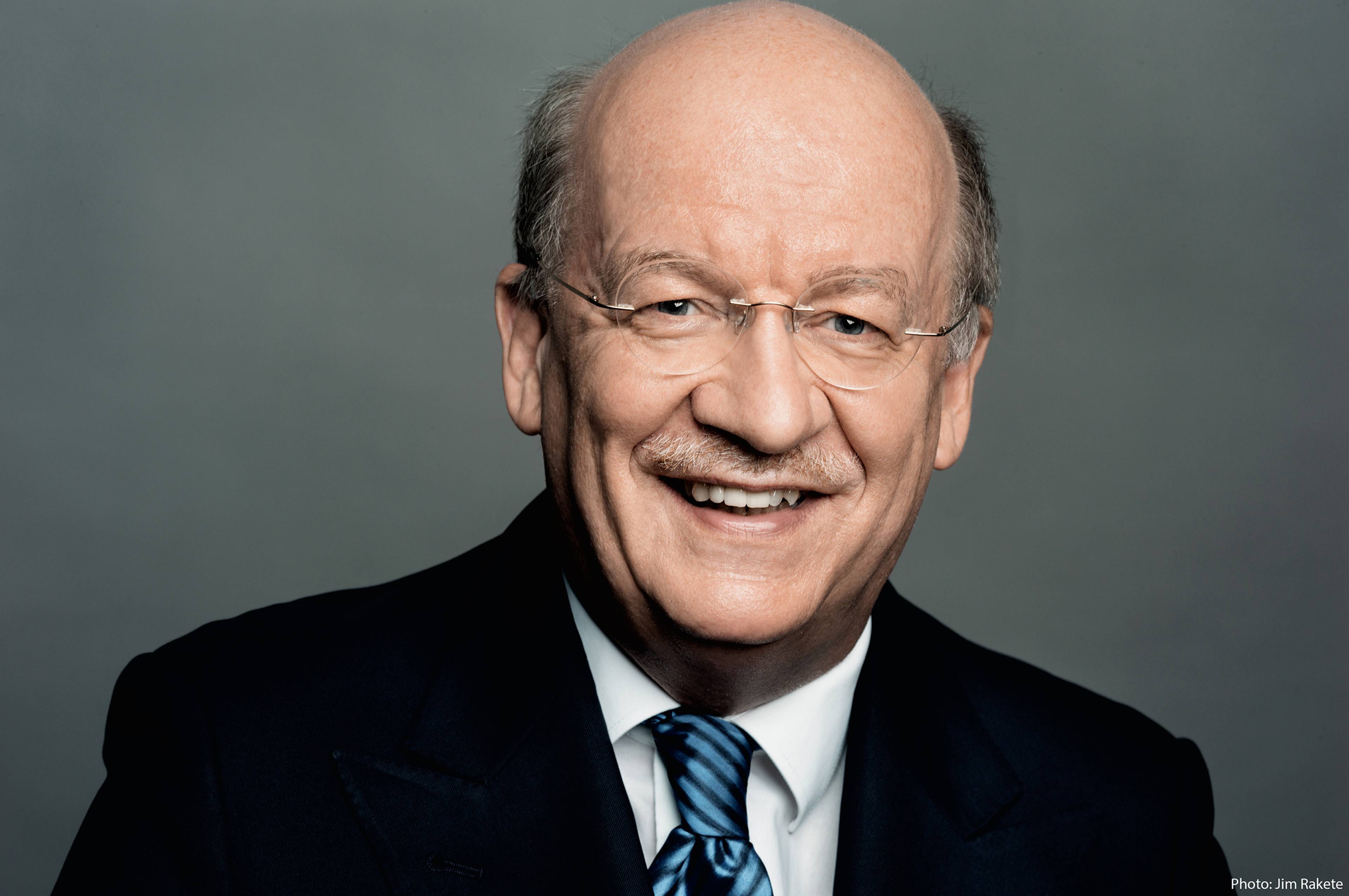 A person smiling while wearing glasses, a dark suit, white shirt, and a striped tie, set against a gray background.