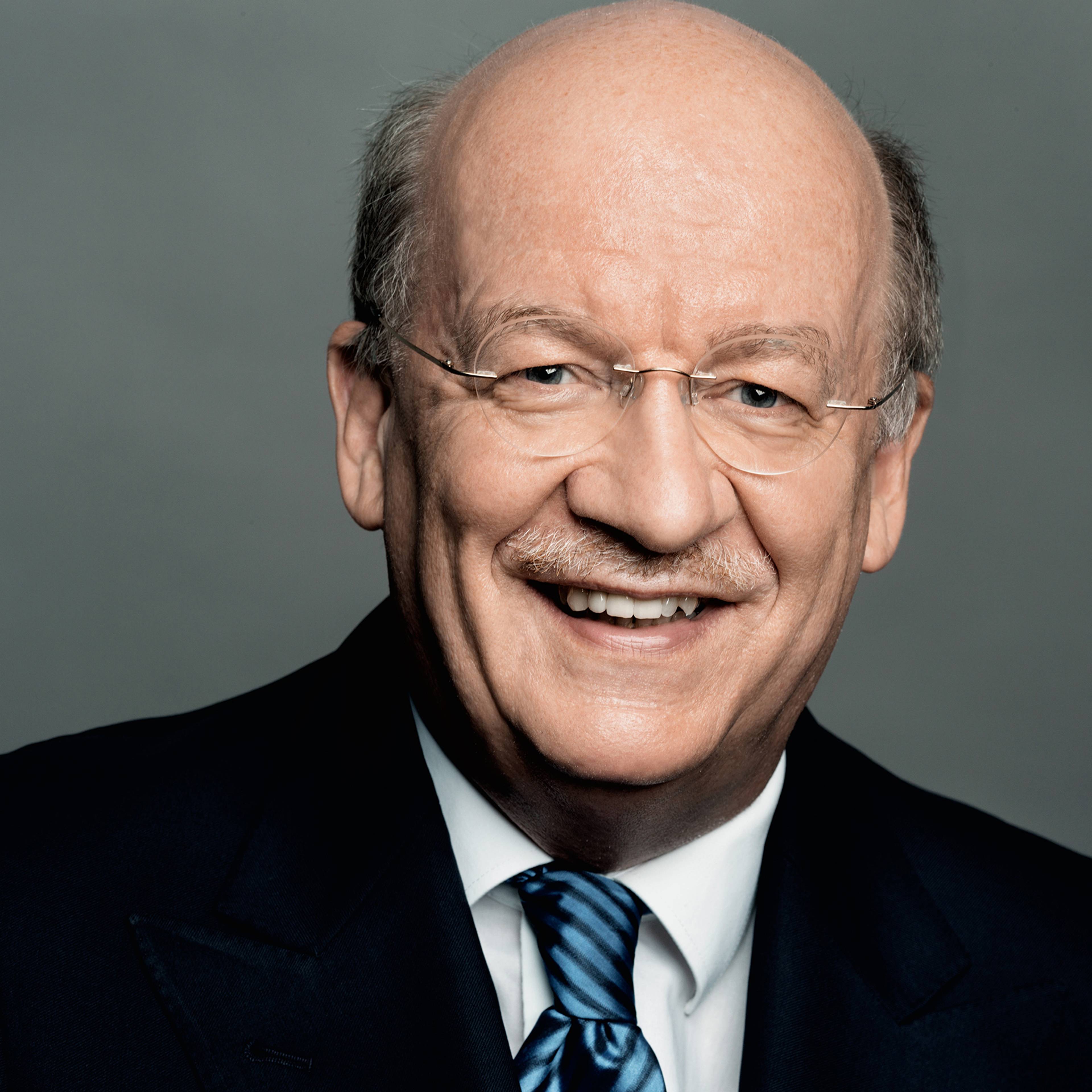 A person smiling while wearing glasses, a dark suit, white shirt, and a striped tie, set against a gray background.