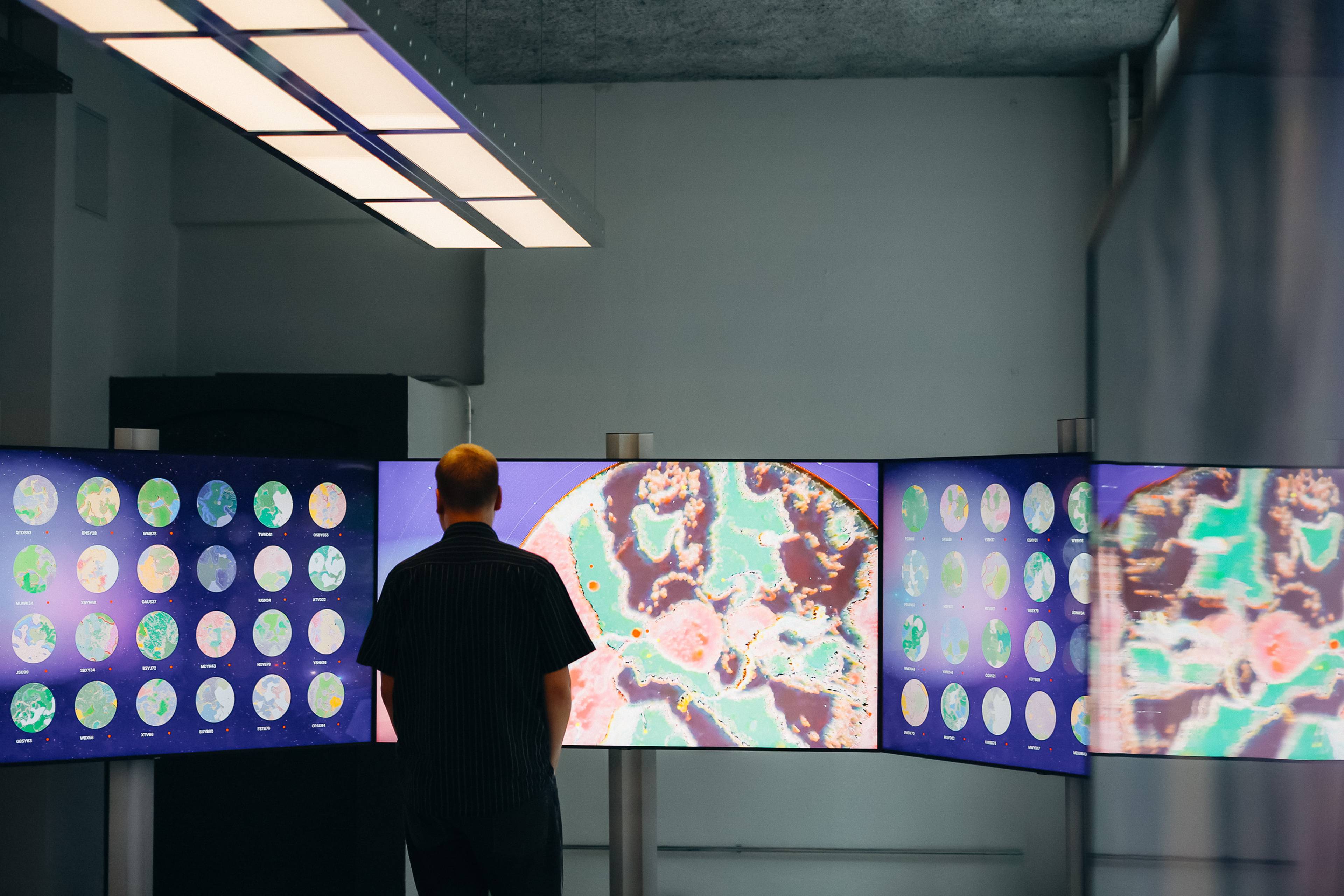 Ein Mann steht mit dem Rücken zur Kamera vor einer gebogenen Anordnung großer Bildschirme, auf denen farbenfrohe digitale Welten zu sehen sind. Die mittleren Bildschirme zeigen Nahaufnahmen abstrakter Landschaften in Pink, Grün, Blau und Weiß, während auf den seitlichen Bildschirmen mehrere kreisförmige Weltansichten dargestellt sind.