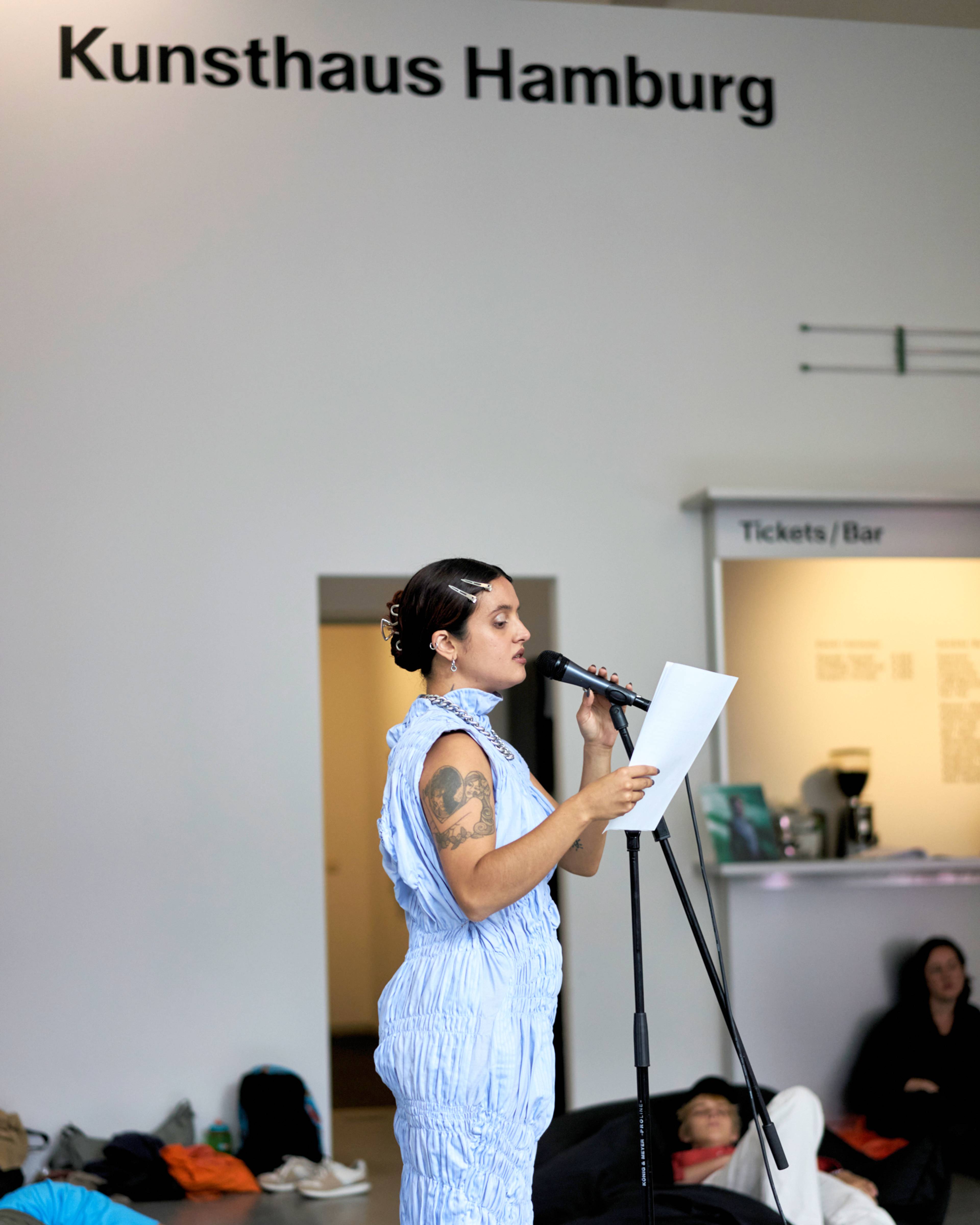 Luïza Luz reading from a paper at a microphone in Kunsthaus Hamburg. Other people are seated on the floor, and a sign for Tickets/Bar is visible in the background.