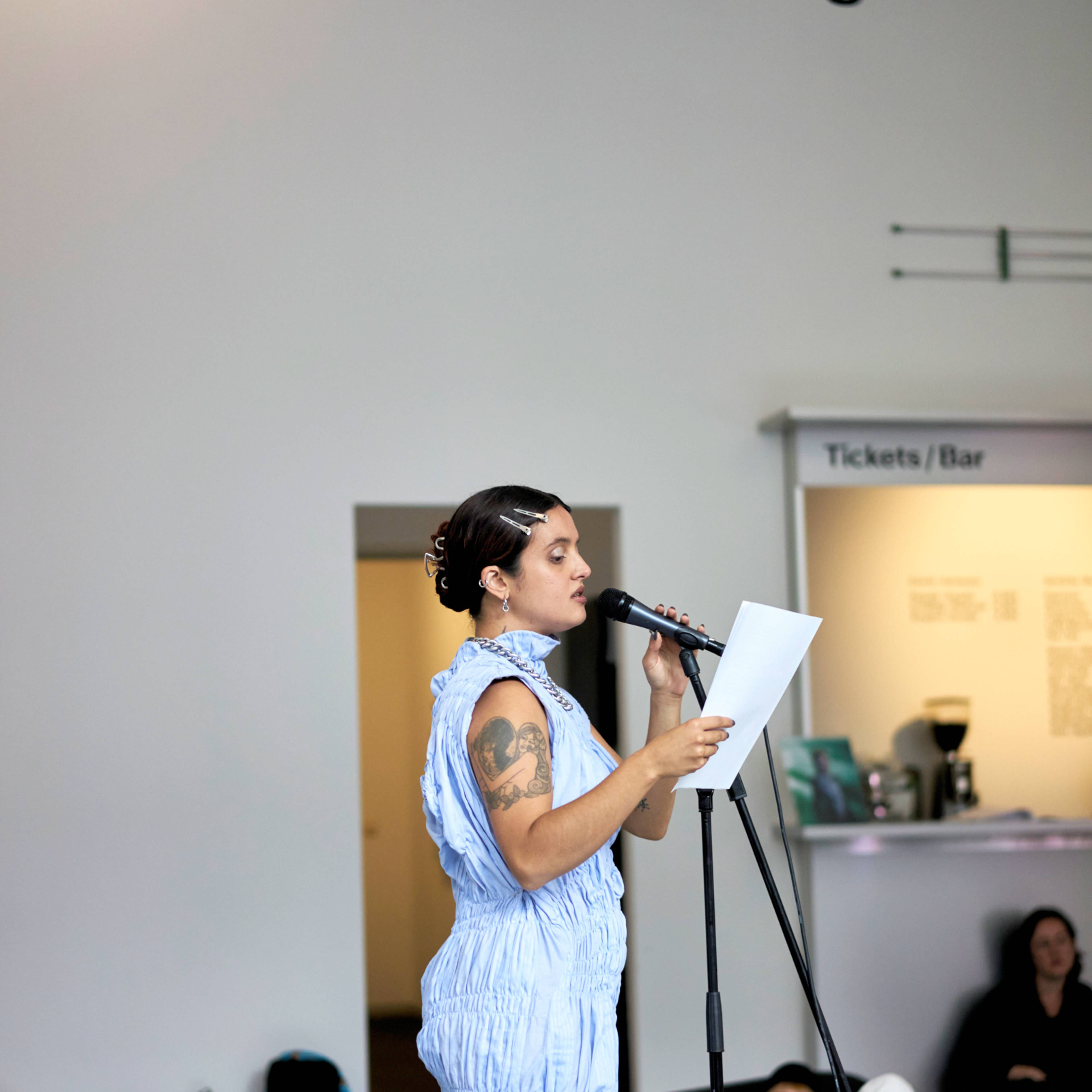 Luïza Luz reading from a paper at a microphone in Kunsthaus Hamburg. Other people are seated on the floor, and a sign for Tickets/Bar is visible in the background.
