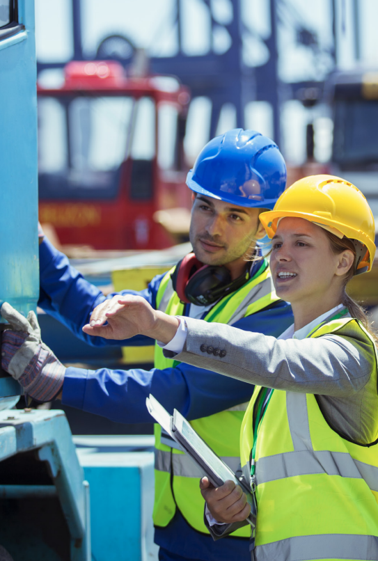 Two industrial workers in hard hats and hi-vis vests inspecting equipment with a tablet at a manufacturing site