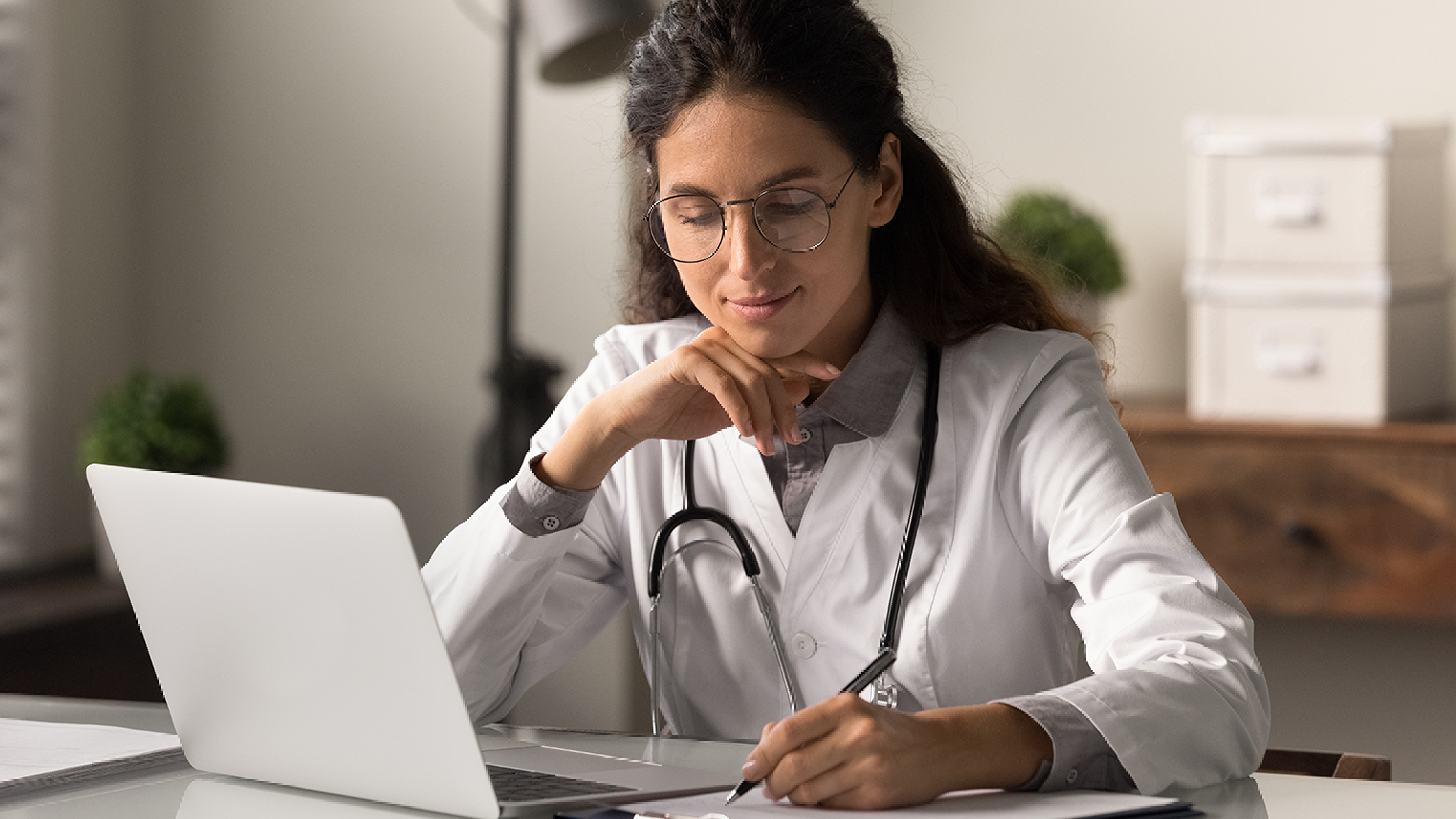 Doctor in a white coat reviewing information on a laptop and taking notes in a healthcare office