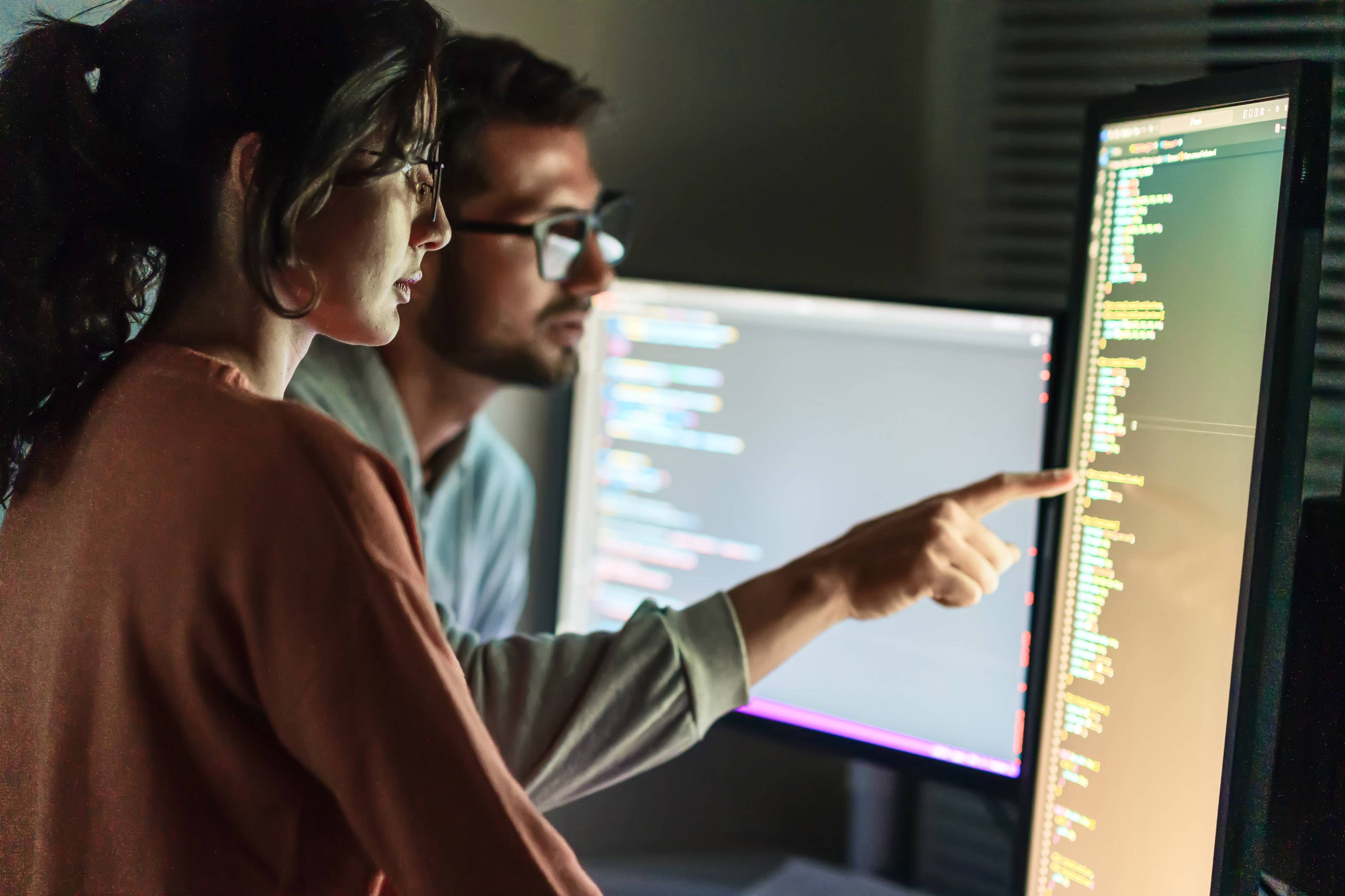 Two IT engineers reviewing code on a large monitor in a network operations centre