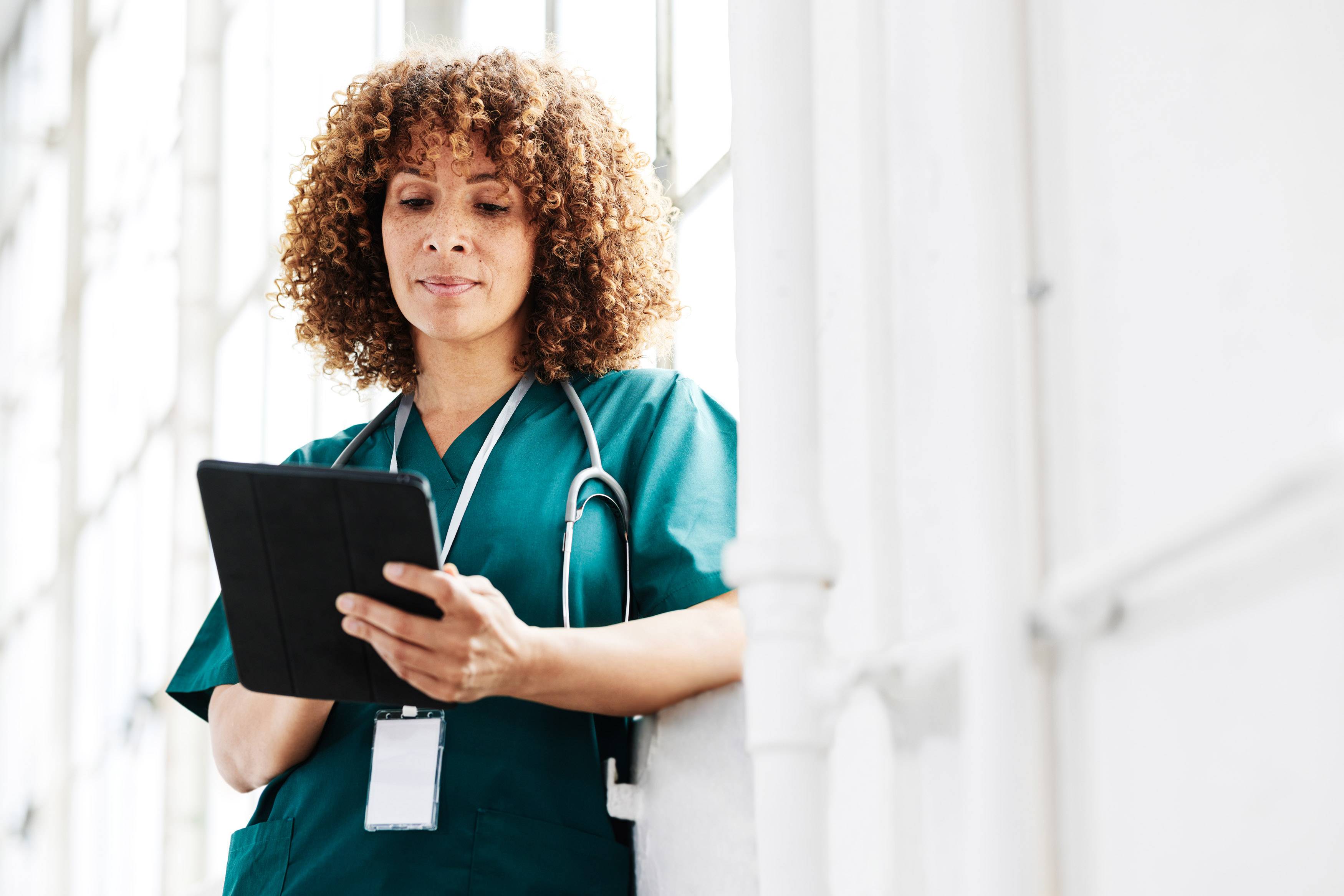 Nurse in scrubs reviewing patient data on a tablet in a bright hospital corridor