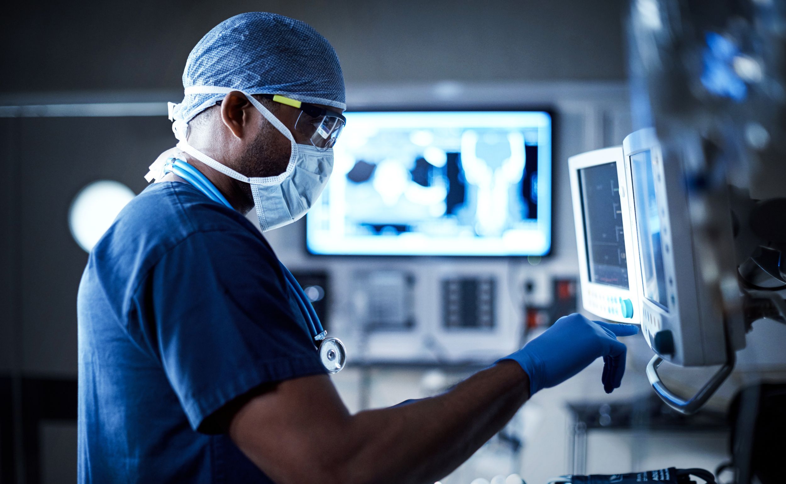 Clinician in surgical scrubs operating a connected patient monitor beside medical imaging screens in a hospital theatre