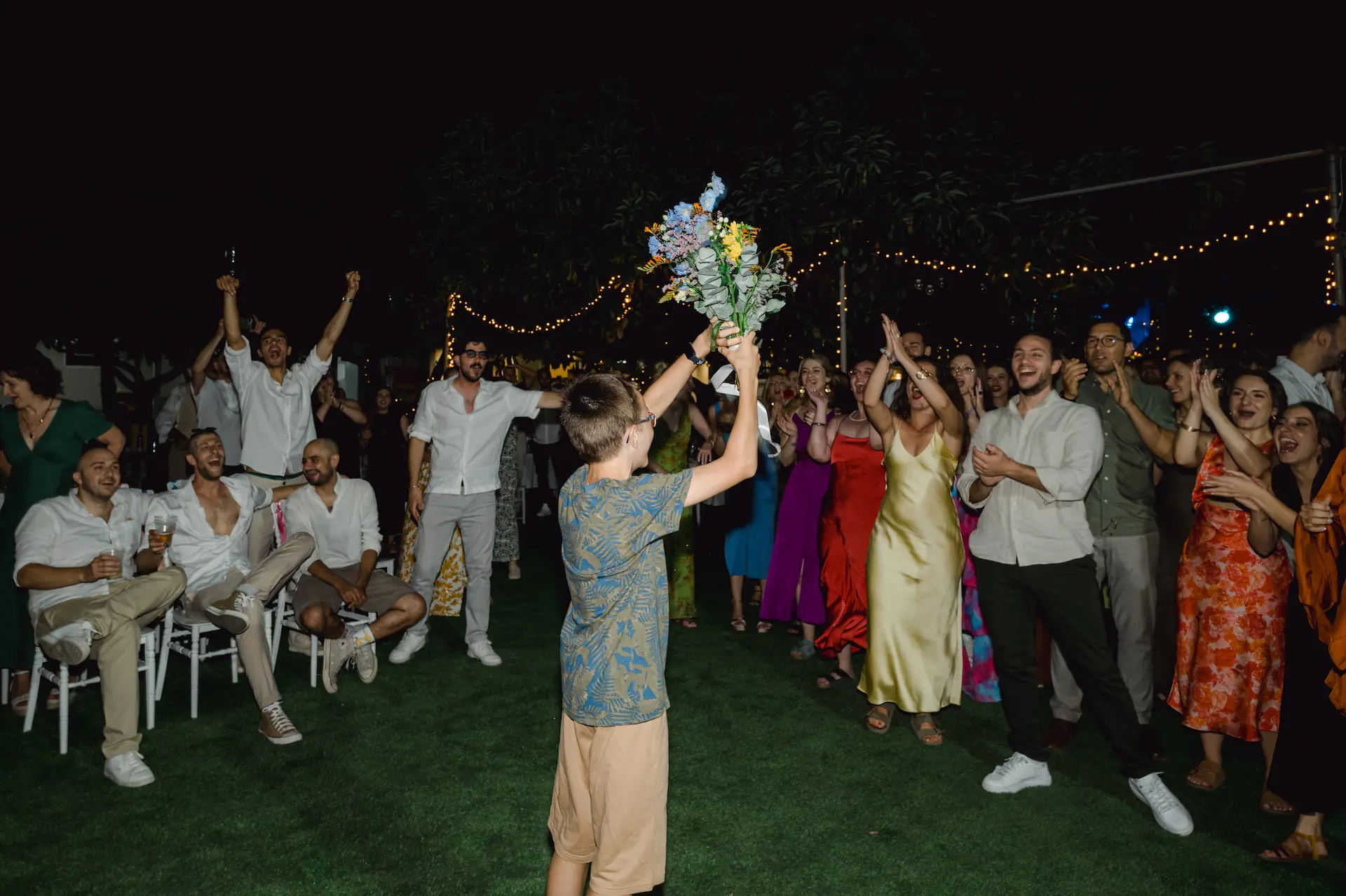 A young boy holding a wedding bouquet while wedding guests cheer him up during the reception.