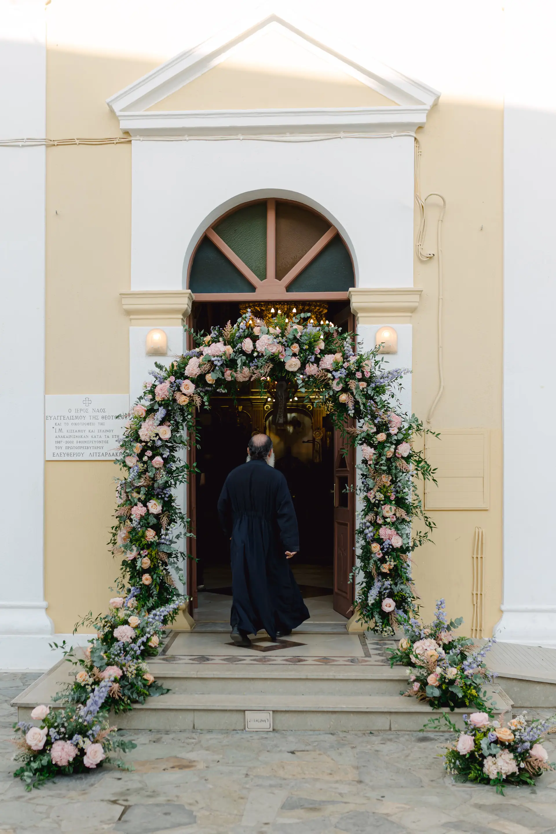 Orthodox priest walking through a floral wedding arch, signaling the beginning of the ceremony in Paleochora