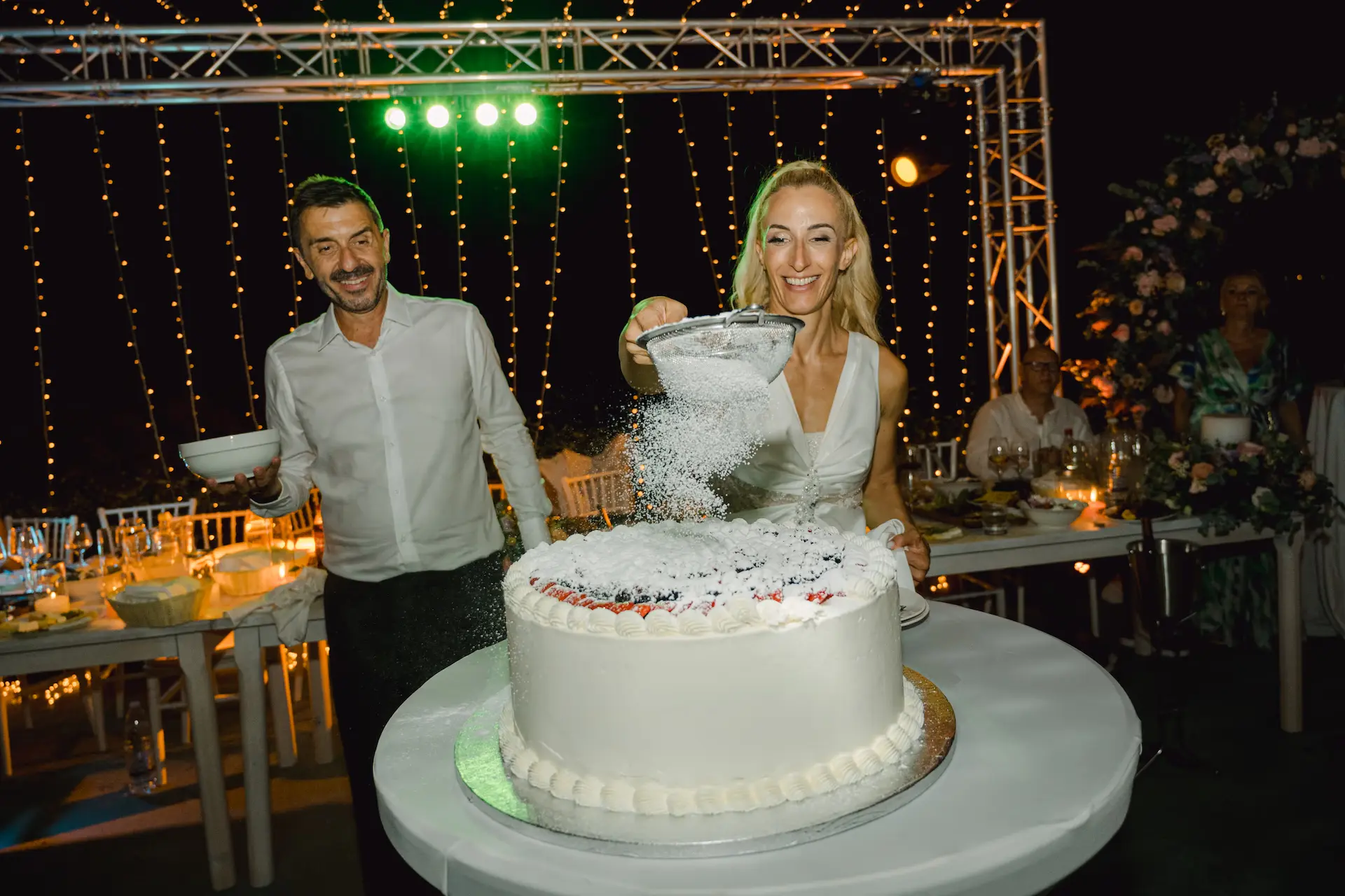 A sweet and candid moment as the bride adds the finishing touch of sugar to her fruit-filled wedding cake 