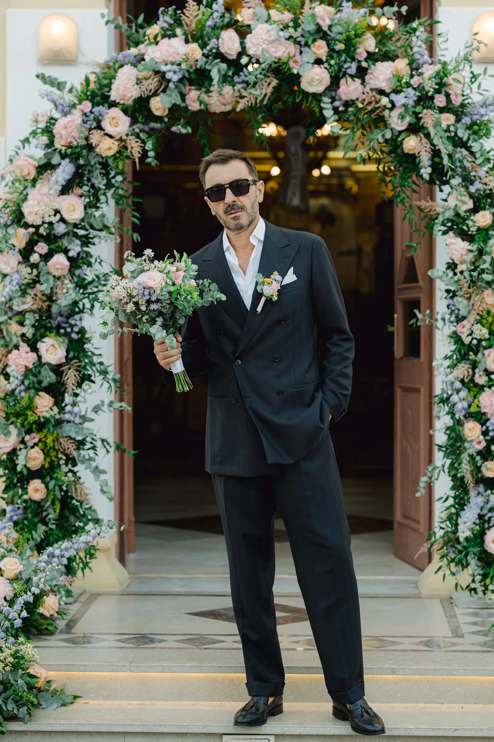 Groom holding a beautiful wedding bouquet while waiting in front of a floral arch at an outdoor church in Paleochora