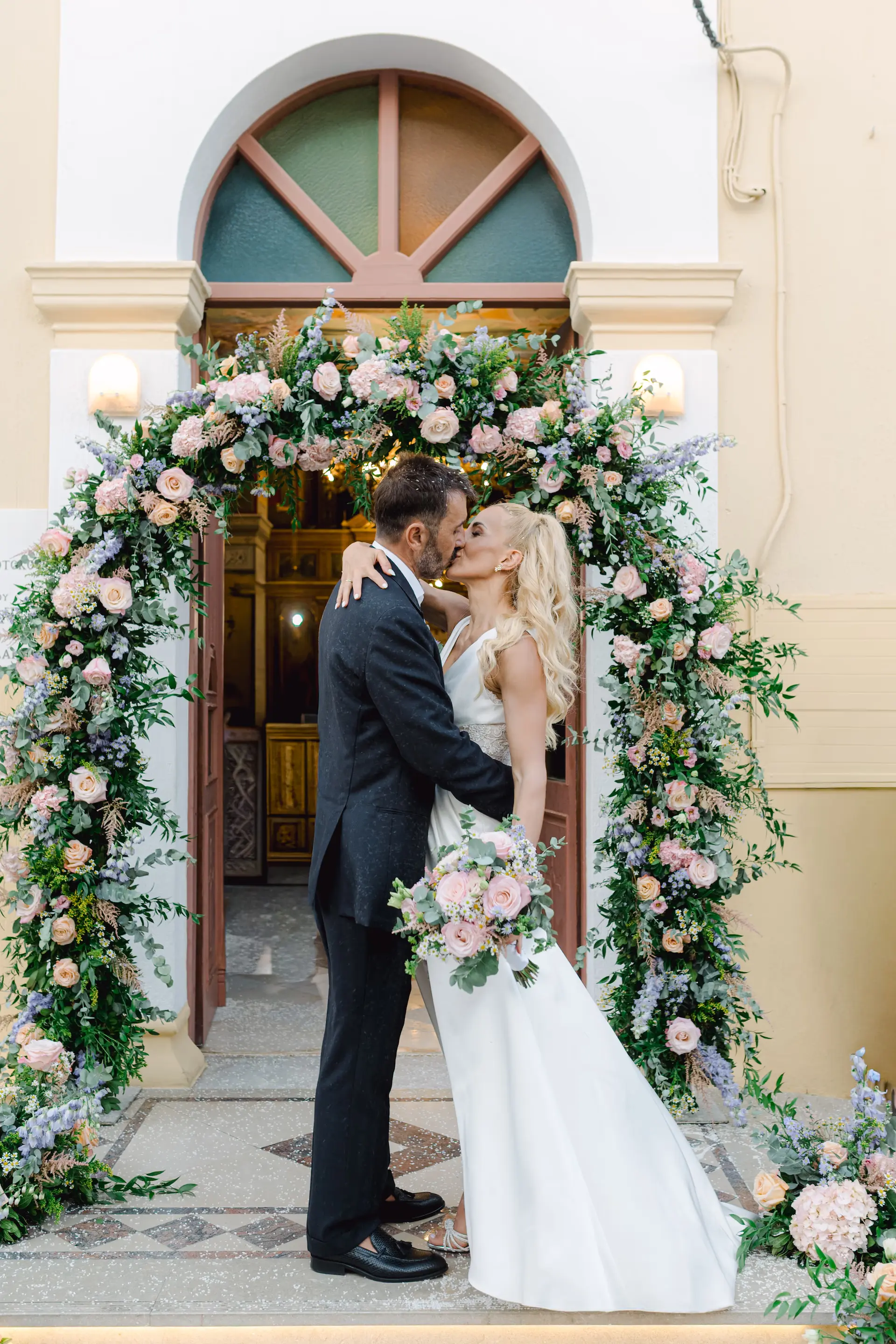 Romantic first kiss of the bride and groom under a stunning white flower arch, capturing a perfect moment of love and celebration.