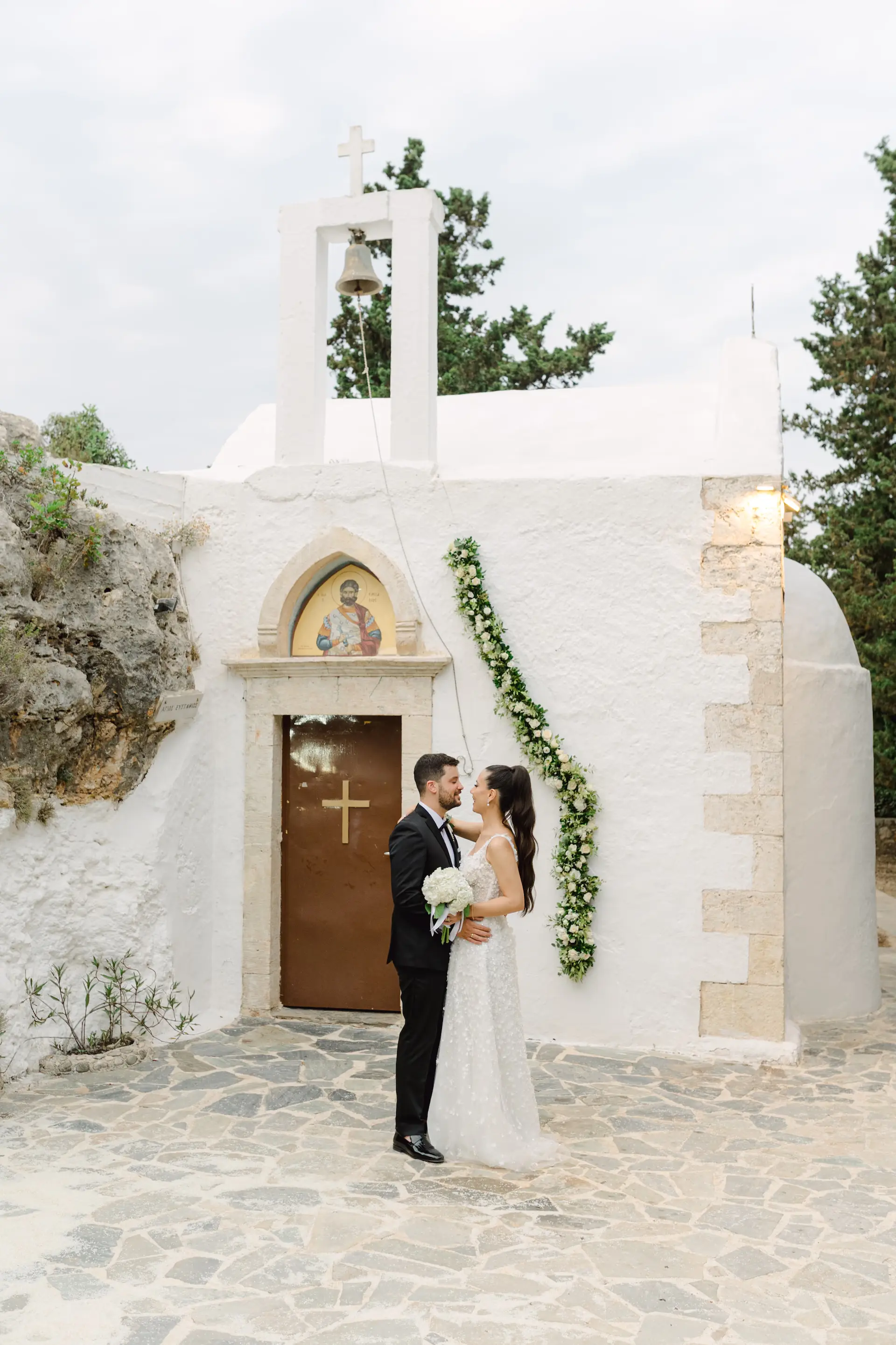 Happy bride and groom laughing together during their wedding portrait session outside a traditional stone church in Chania.