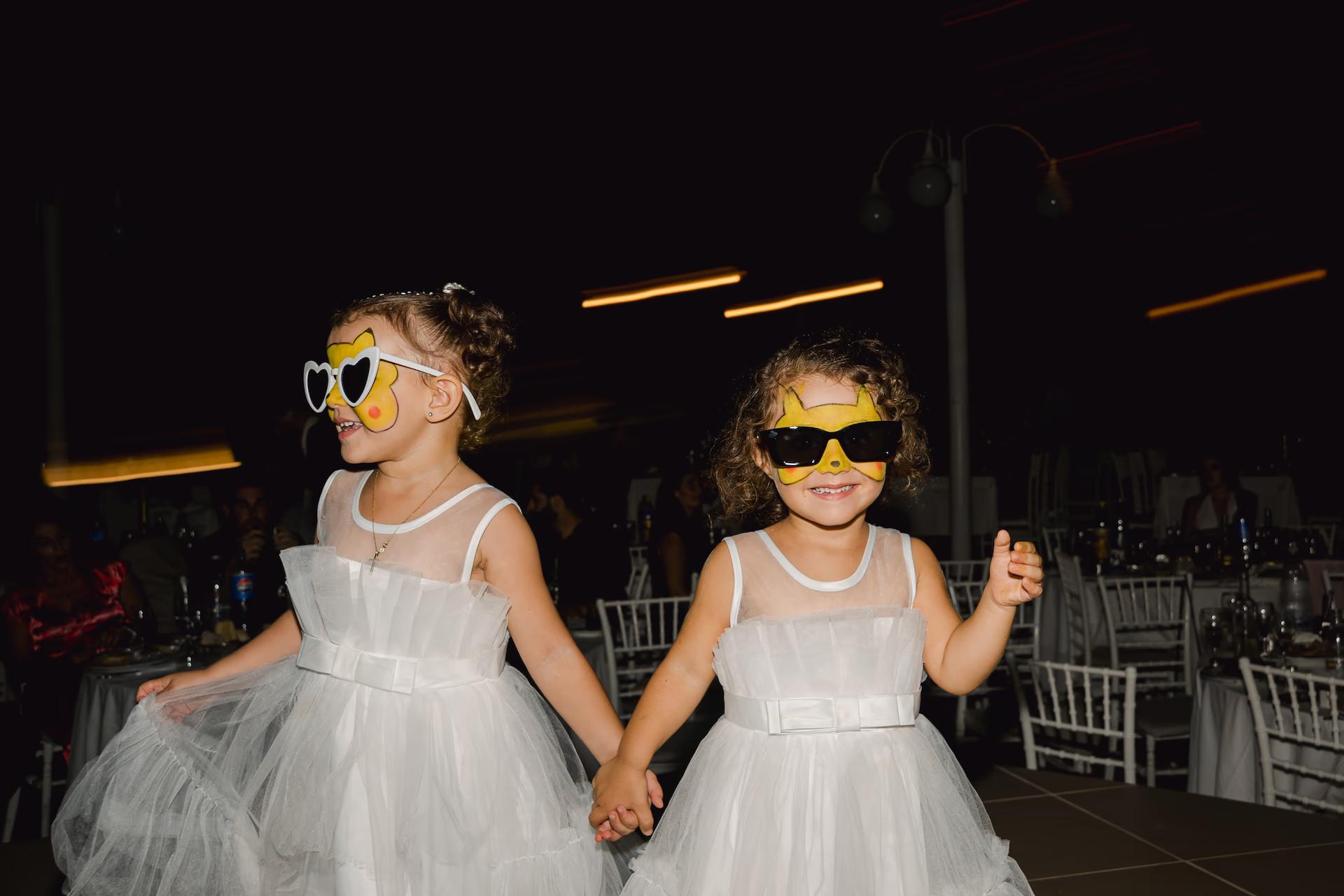 Adorable flower girls in white dresses wearing matching sunglasses, having fun at a wedding reception.