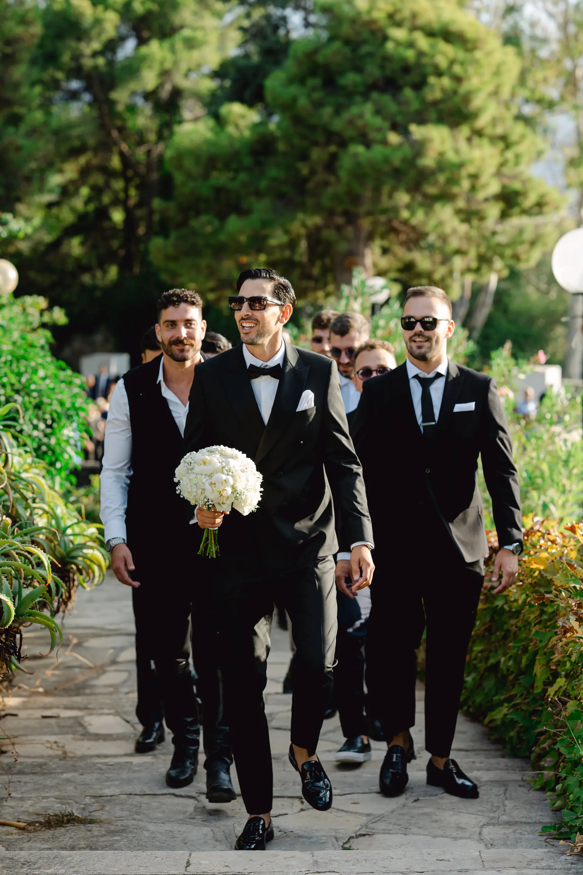 groom and groomsmen in suits walking down a path with flowers