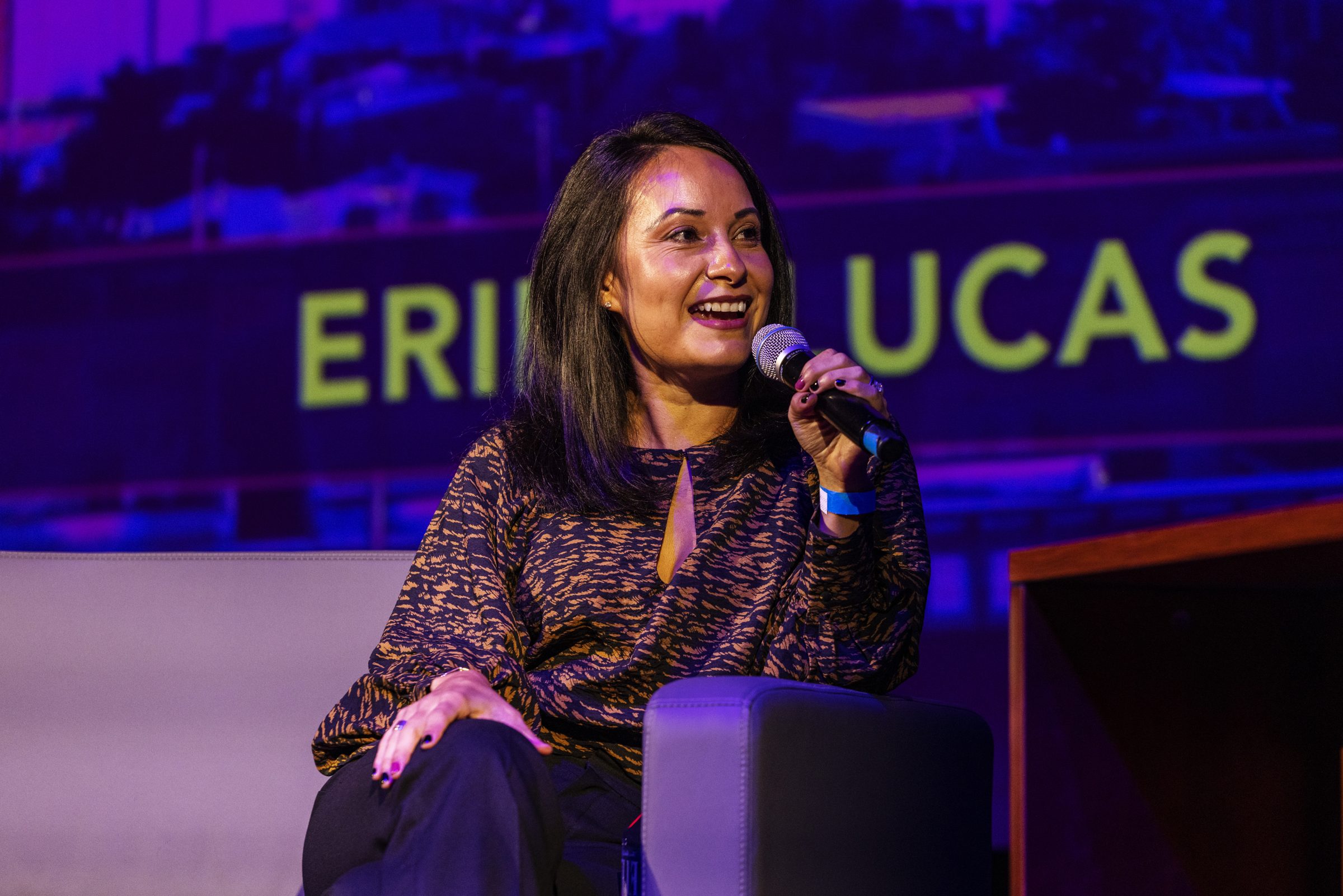A guest smiles and speaks into a microphone, her name projected on the screen behind her.