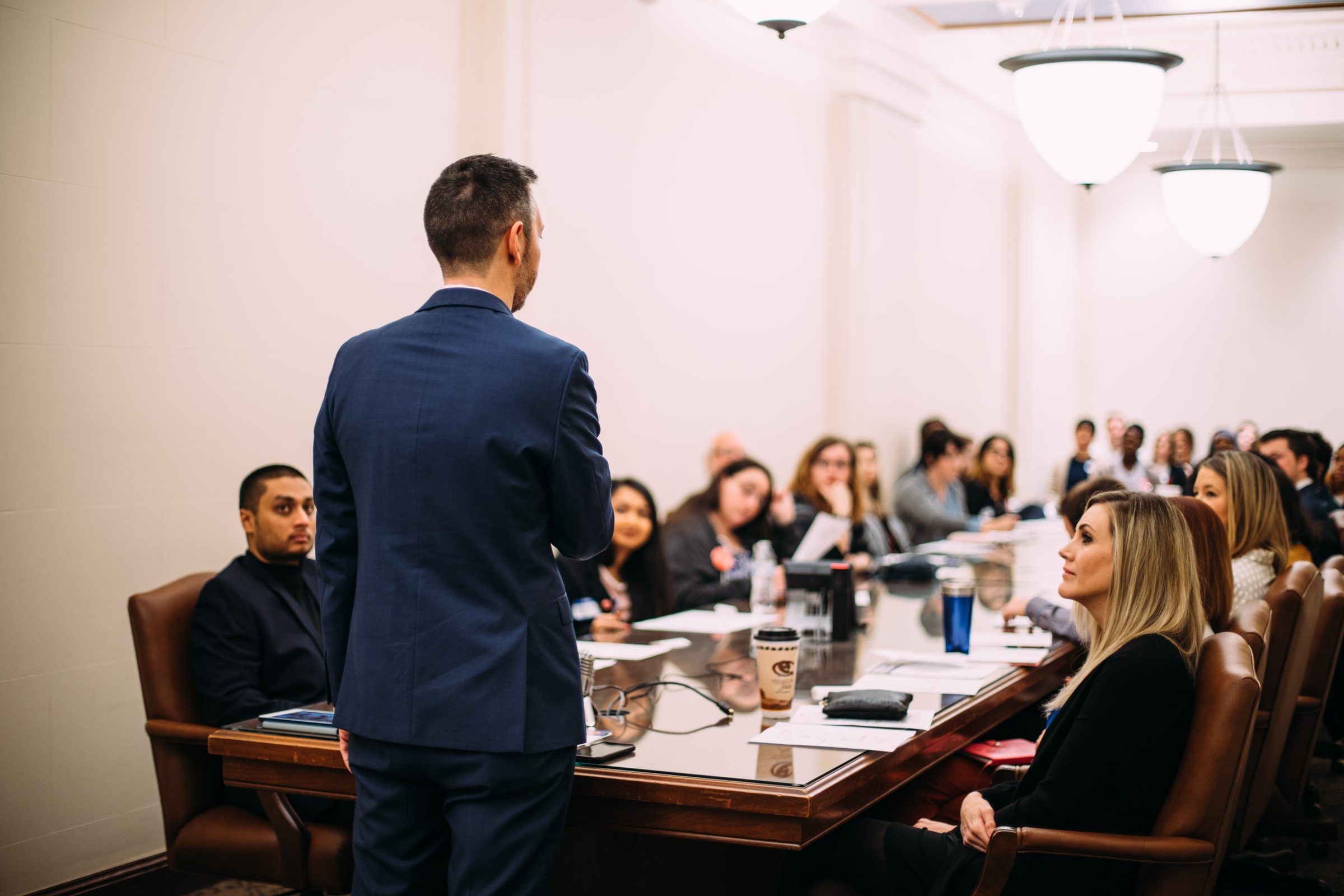 A presenter stands before a full conference table of attendees in a Capitol meeting room.