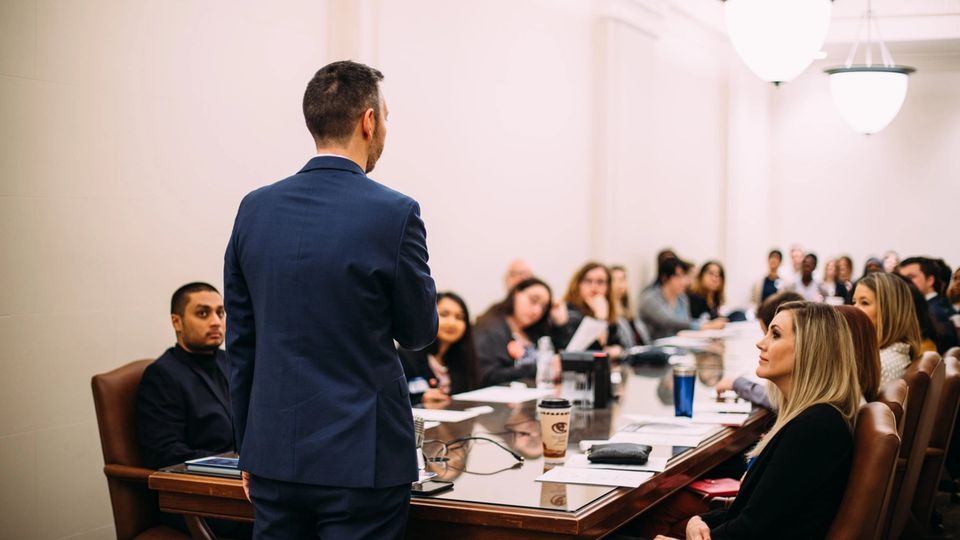 A presenter stands before a full conference table of attendees in a Capitol meeting room.