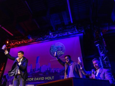 A guest raises a hand toward the audience while seated on stage in front of the skyline backdrop.