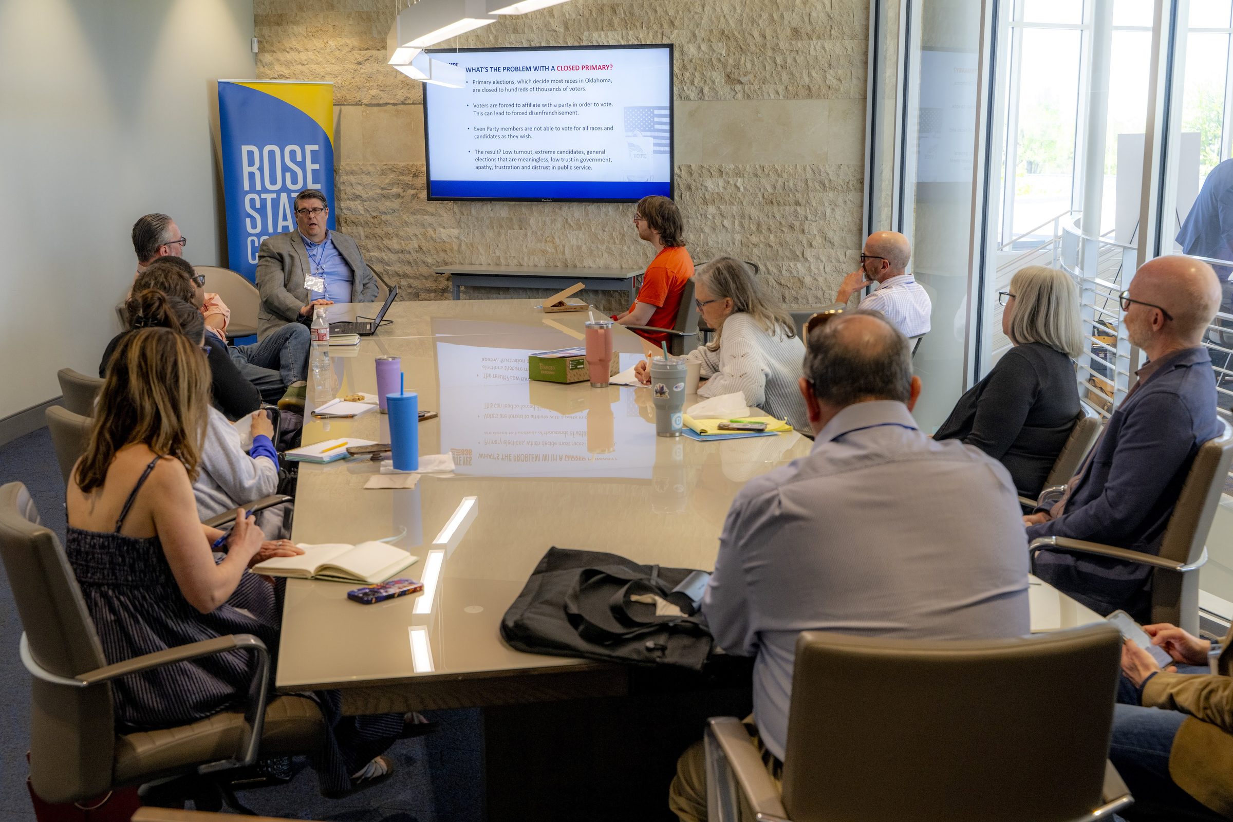 Participants seated around a long conference table listen to a presentation in a breakout room.