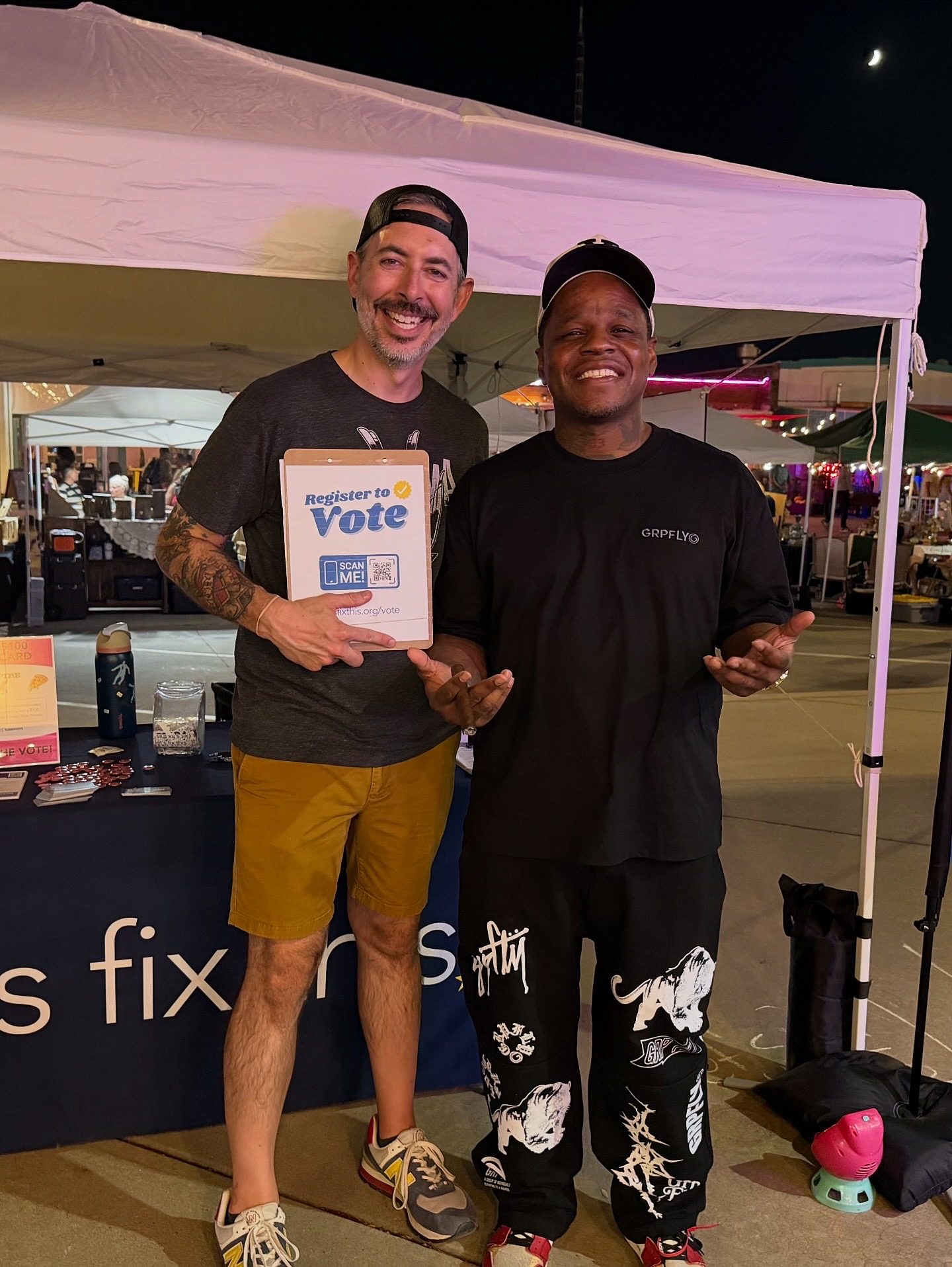 Two men smile beside a Let's Fix This booth at a night market, one holding a Register to Vote clipboard.