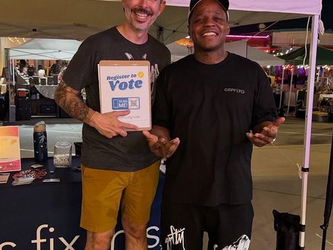 Two men smile beside a Let's Fix This booth at a night market, one holding a Register to Vote clipboard.