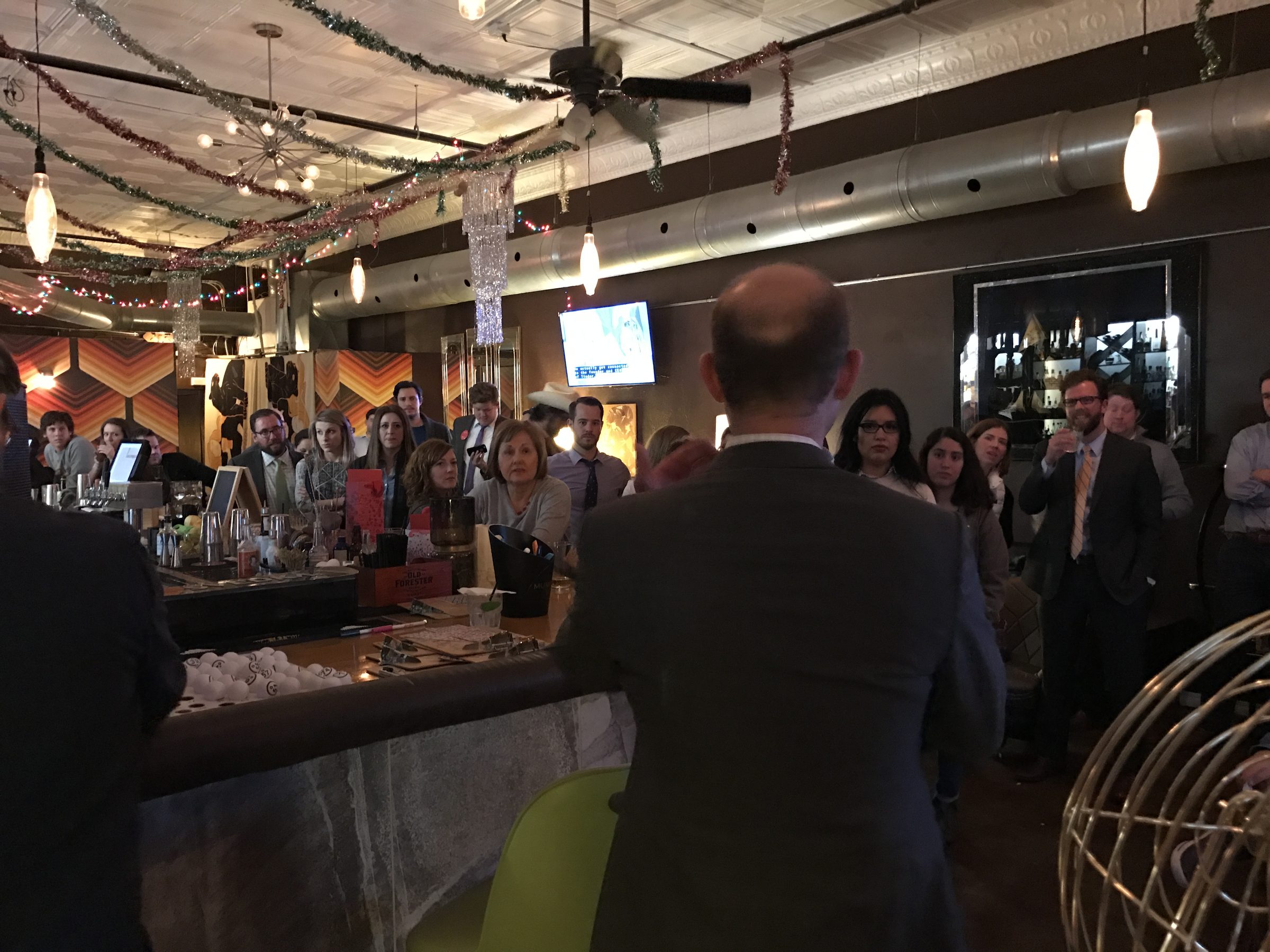 A crowd gathers around a bar to listen to a speaker during an evening event.