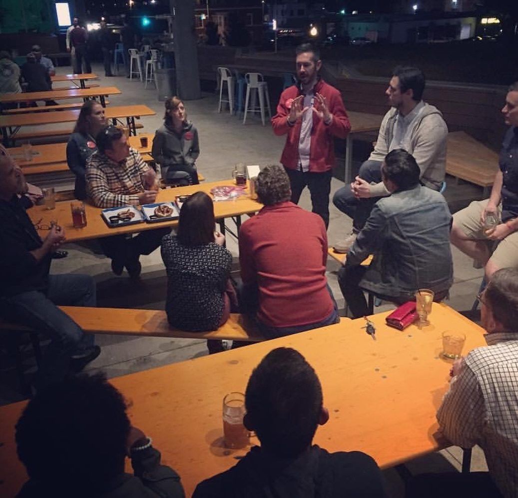 People gather at long tables on a patio while a man speaks to the group.