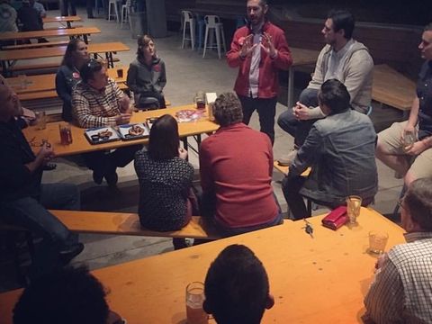 People gather at long tables on a patio while a man speaks to the group.