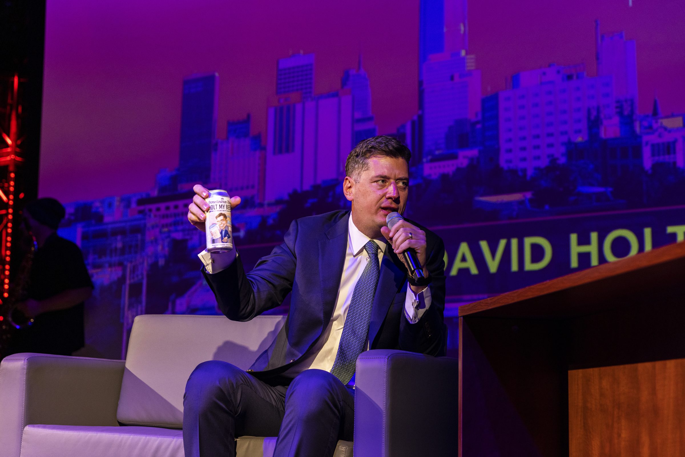 A guest holds up a small cup while talking with the host on stage.