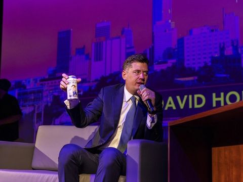 A guest holds up a small cup while talking with the host on stage.