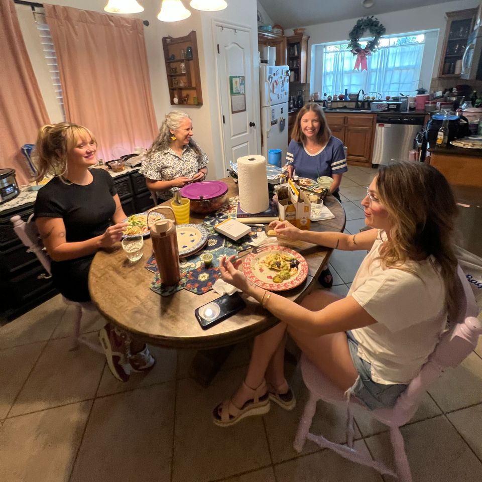 Four women share a potluck dinner around a kitchen table.