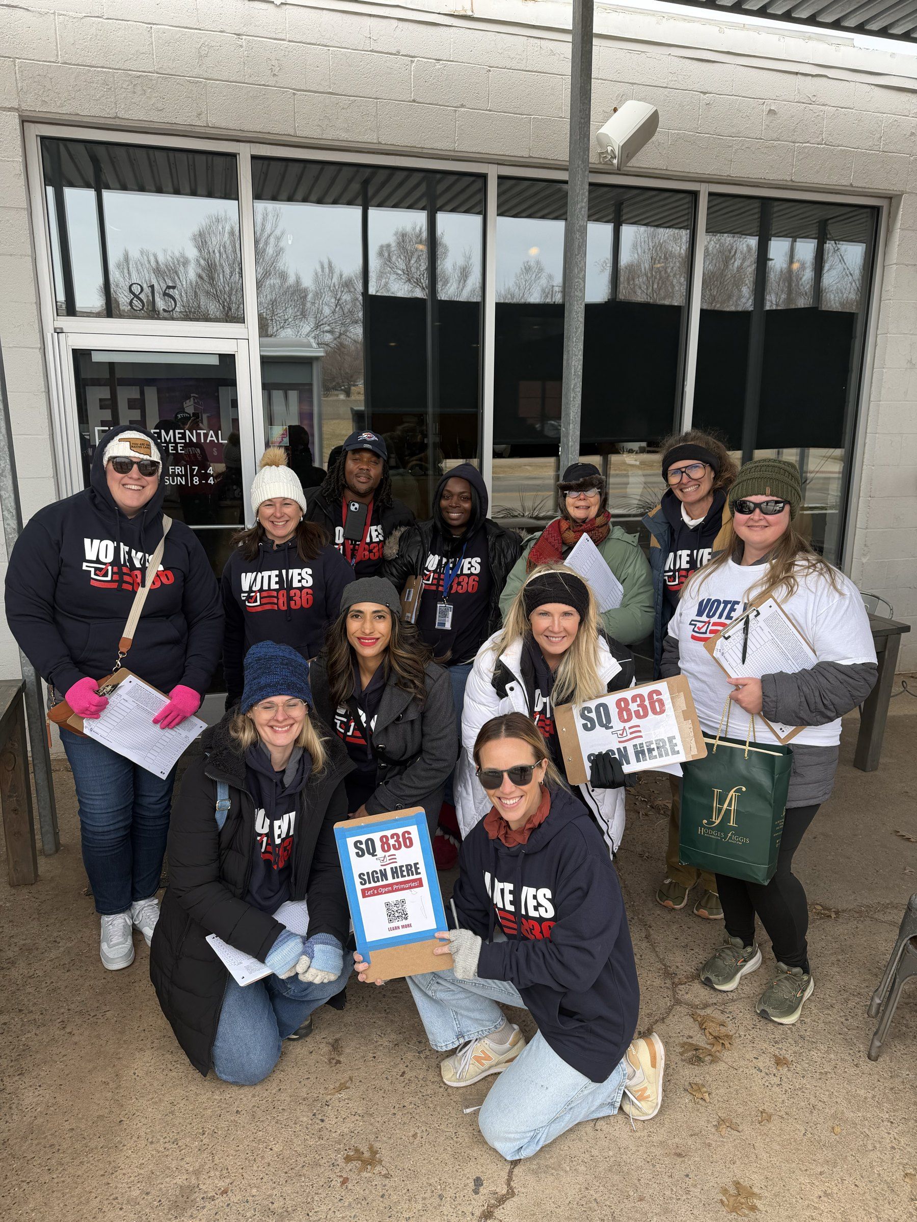 Volunteers in Vote Yes 836 hoodies hold clipboards and “SQ 836 Sign Here” signs outside a storefront during the SQ 836 signature drive.