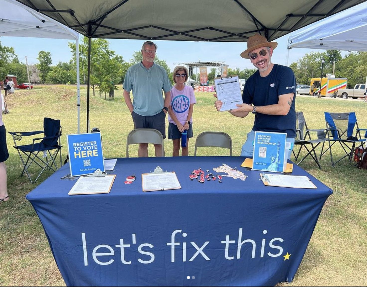 Three volunteers smile behind a Let's Fix This voter registration table set up in a park.