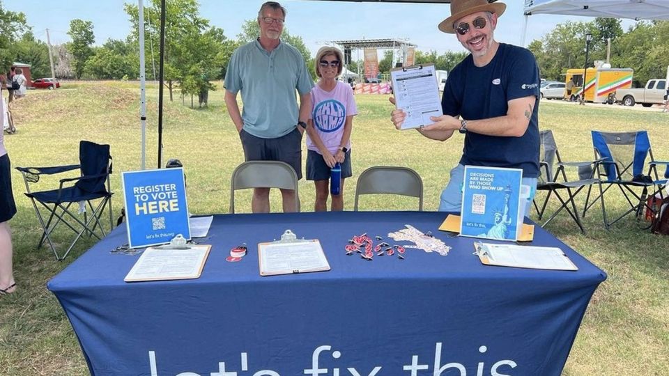 Three volunteers smile behind a Let's Fix This voter registration table set up in a park.