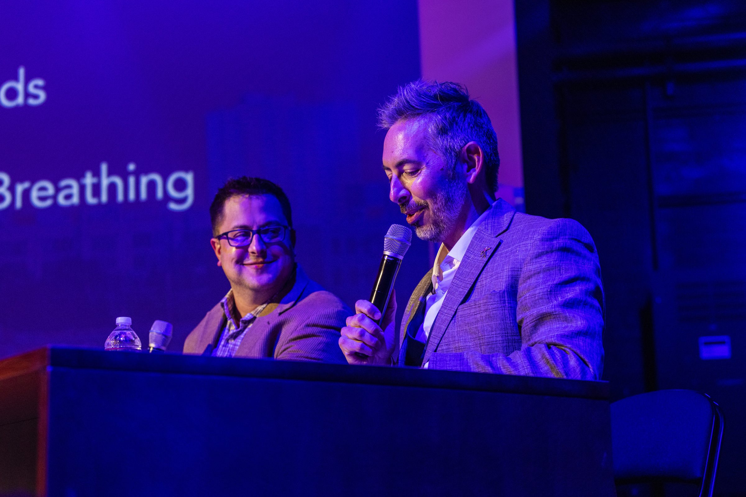 A host and a guest laugh while talking at the interview desk on stage.