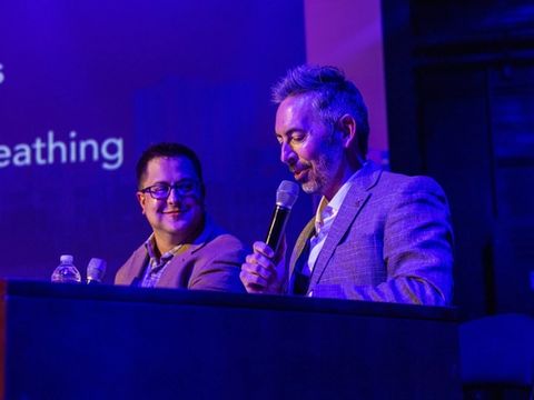 A host and a guest laugh while talking at the interview desk on stage.