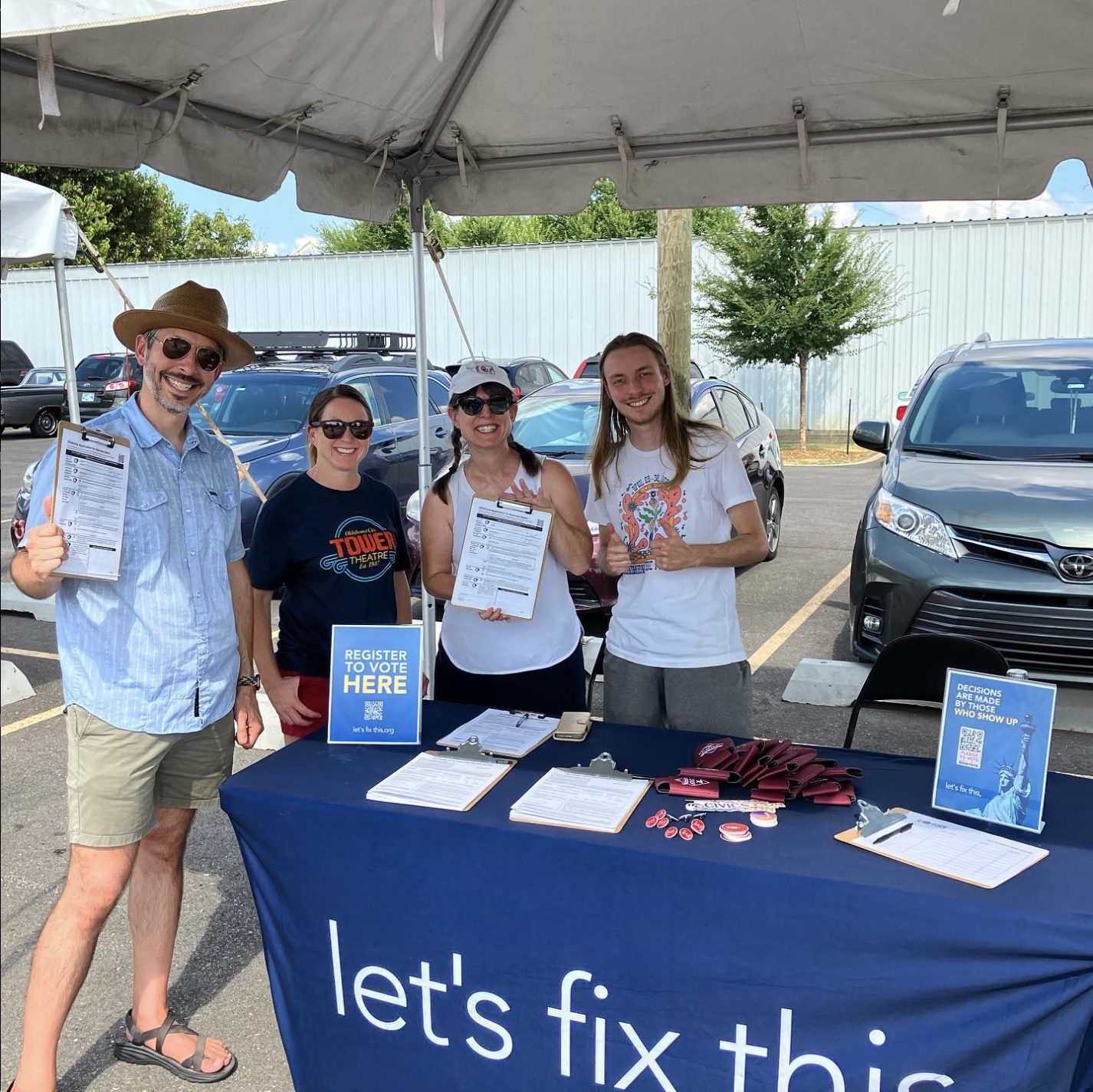 Four smiling volunteers hold voter registration forms behind a Let's Fix This table under a tent.