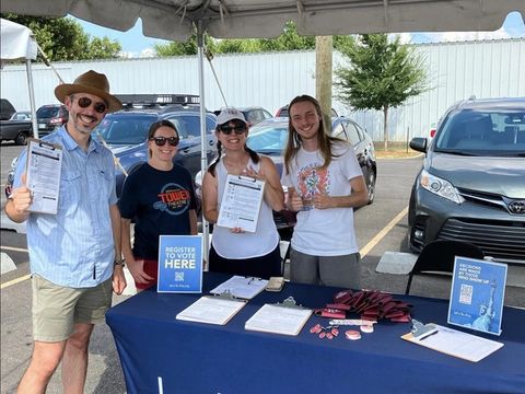 Four smiling volunteers hold voter registration forms behind a Let's Fix This table under a tent.