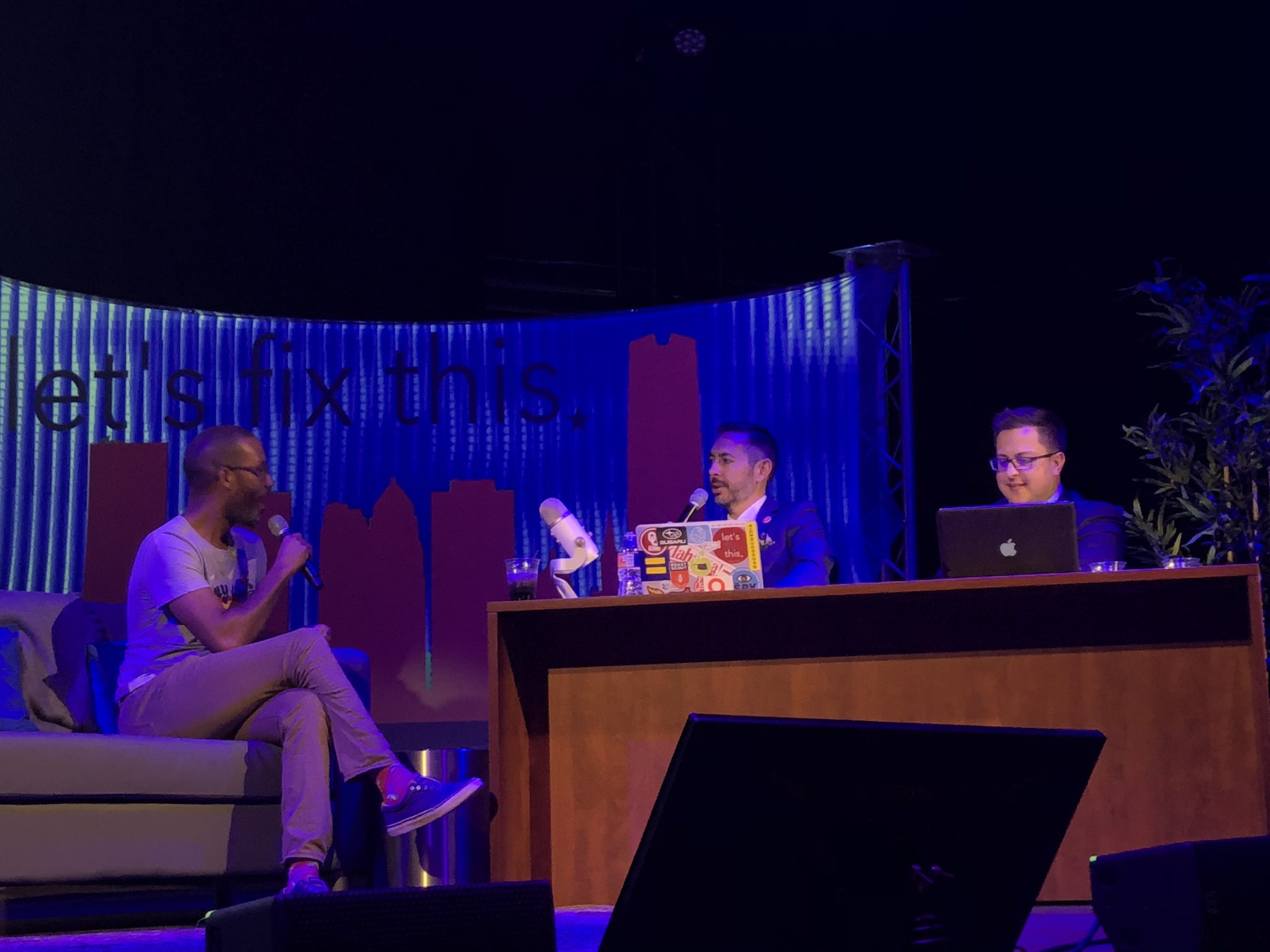 A guest on a couch talks with two hosts at a news desk in front of a glowing Let’s Fix This backdrop.