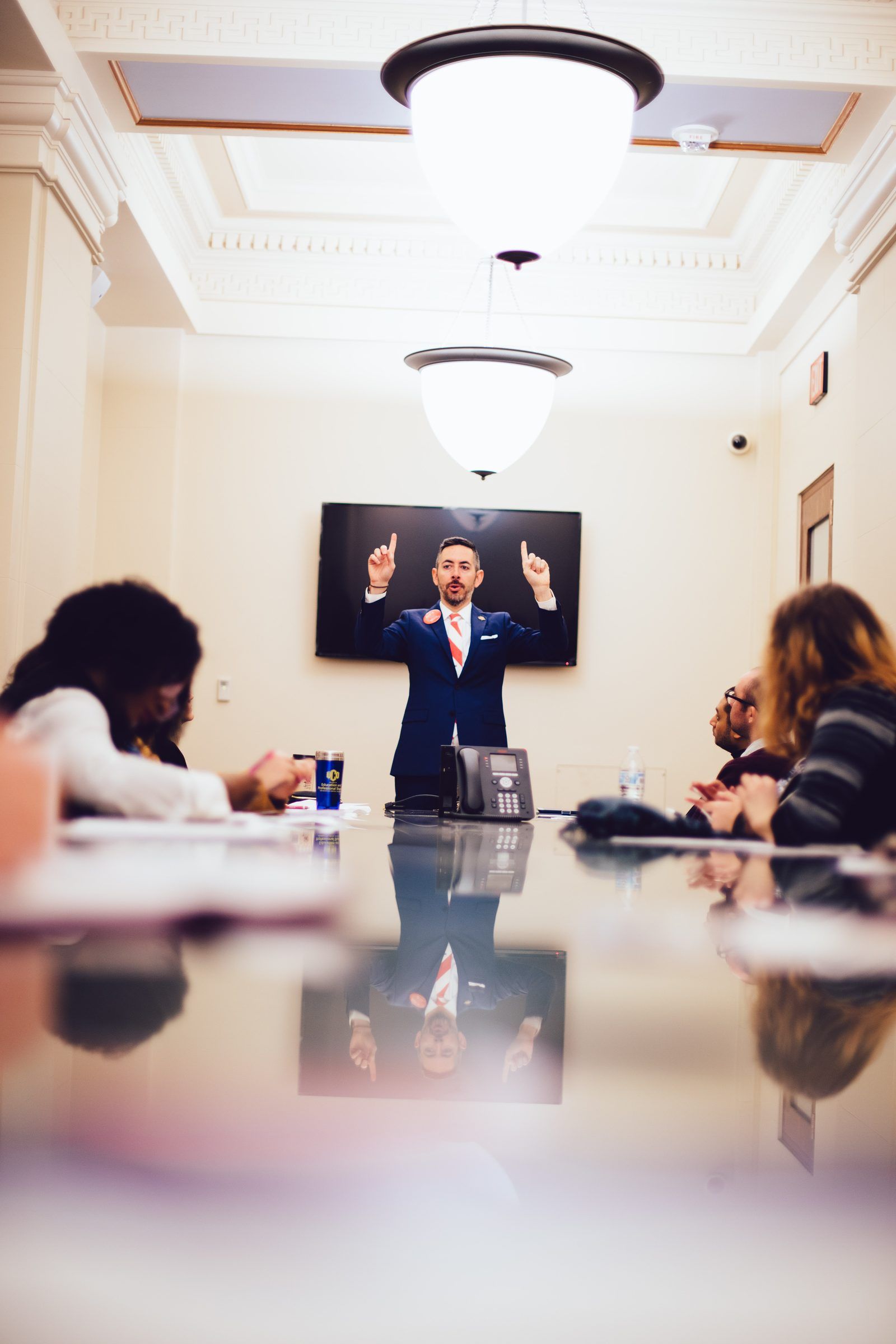 A speaker gestures at the head of a long conference table as attendees listen.